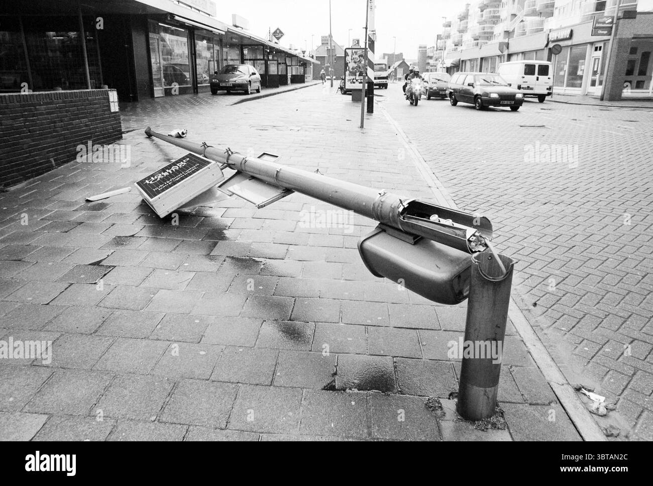 Sturmstrand + Dorf Zandvoort Zandvoort, Whizgle News, Dutch Desk, Niederlande, 1950 - 2000 auf 31-03-1994. Das Bild enthält diese Themen. Die Szene zeigt eine Straße, die ein Gefühl von Vernachlässigung und Nachwirkungen vermittelt. Ein Straßenlaternenmast liegt auf der Seite, was darauf hindeutet, dass er beschädigt wurde, möglicherweise durch Unwetter oder einen Unfall. Die Oberfläche unter dem Stab besteht aus quadratischen, dunklen Fliesen, die Anzeichen von Feuchtigkeit und möglicherweise Regen reflektieren. Um den abgestürzten Pol herum befinden sich Gebäude entlang der Straße, deren Fassaden eine Mischung aus gedämpften Farben, überwiegend Grau- und verwitterten Tönen, aufweisen Stockfoto