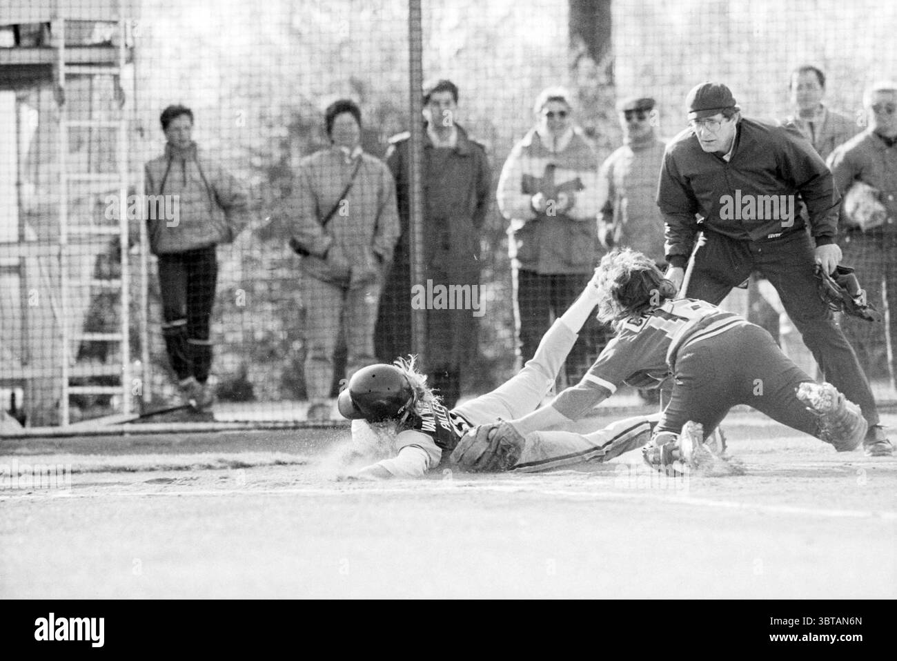 Terrasvogels - HCAW Softball, Whizgle News, Dutch Desk, Niederlande, 1950 - 2000 am 24-04-1988. Die Abbildung zeigt diese Themen. In einer dynamischen Outdoor-Umgebung spielen zwei Spieler während eines Baseballspiels einen angespannten Moment. Der Spieler, der in die Startplatte gleitet, trägt einen dunklen Helm und ein Trikot, im Kontrast zu der leichteren Uniform des Fingers, der in einer hockenden Haltung positioniert ist und sichtbar versucht, den Schiebespieler zu markieren. Der Boden unter ihnen ist eine Mischung aus Schmutz und Gras, die Anzeichen von Abnutzung durch das Spiel zeigt. Um das Geschehen herum beobachten mehrere Zuschauer aufmerksam hinter einem Ofen Stockfoto