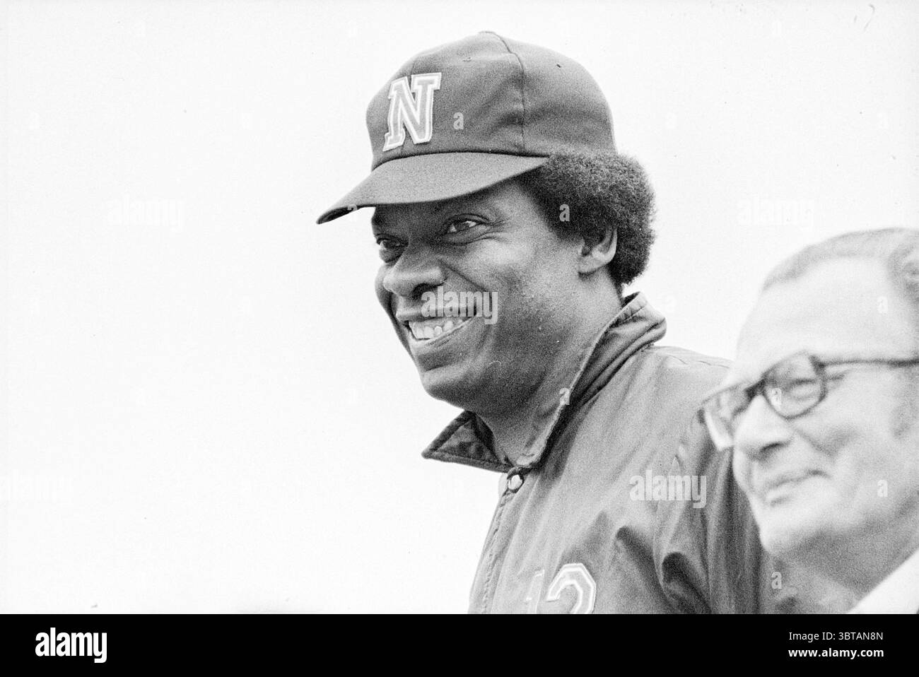 Head Dutch Baseball Team (Namen siehe Titelseiten) Baseball, Whizgle News, Dutch Desk, Niederlande, 1950 - 2000 am 22.07.1975. Diese Themen sind in der Abbildung dargestellt. Die Szene zeigt zwei Personen, die beide Ausdruck von Freude und Engagement ausstrahlen. Eine Person trägt eine dunkle Mütze mit einem prominenten Buchstaben „N“ und hat ein breites, warmes Lächeln, das ihre Begeisterung zum Ausdruck bringt. Die Person hat ein unterscheidbares, eng gewickeltes Afro, das im Kontrast zu den hellen Tönen ihrer Jacke steht, die sportlich wirkt. Neben diesem Individuum steht eine zweite Figur, die kurze Haare hat und trägt Stockfoto