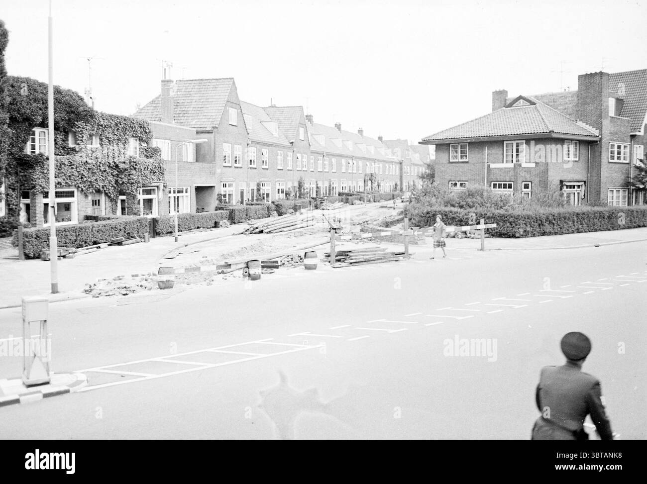 Straßenbauecke Kleverlaan/Engelszstraat Haarlem Kleverlaan the Netherlands, Whizgle News, Dutch Desk, Niederlande, 1950 - 2000 im Jahr 1965. Das Bild enthält diese Themen. Die Szene zeigt eine Wohnstraße aus der Ferne, die eine Mischung aus architektonischen Stilen zeigt, die hauptsächlich durch zweistöckige Backsteingebäude geprägt sind. Die Farbpalette neigt zu gedämpften Grautönen und tiefen Brauntönen, wobei einige Strukturen einen Hauch von Grün zeigen, während sich Efeu an den Wänden anschmiegt. Im Vordergrund ist eine Straße sichtbar, leicht abgenutzt und durch weiße Linien gekennzeichnet. Sie wird von einem erhöhten Rand begrenzt Stockfoto