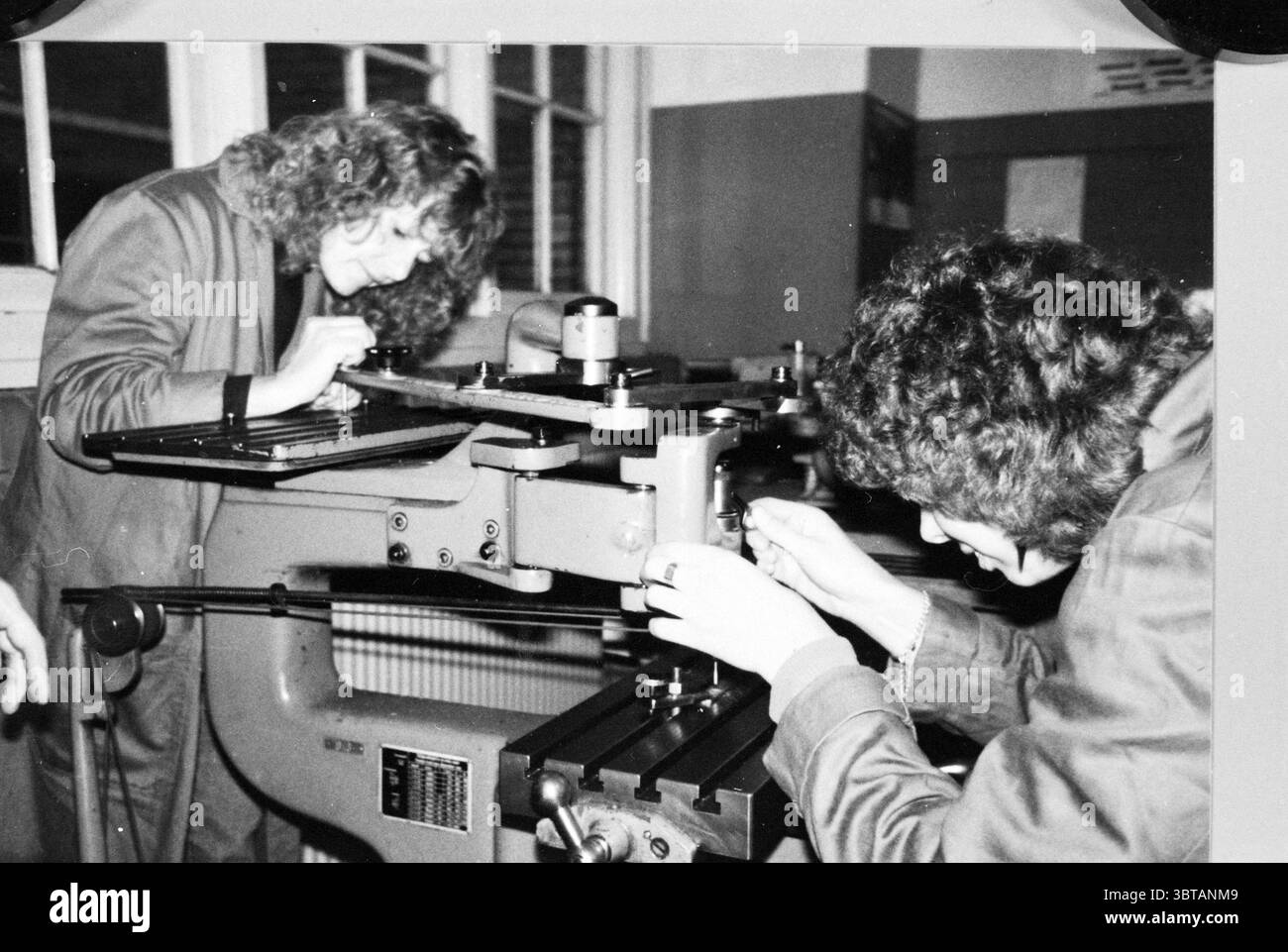 Präzisionsarbeit an Maschine., Whizgle News, Dutch Desk, Niederlande, 1950 - 2000. Diese Themen werden in der Abbildung angezeigt. Die Szene zeigt zwei Personen, die sich intensiv auf eine große, industrielle Maschine konzentrieren, wahrscheinlich ein Fräs- oder Bearbeitungswerkzeug. Beide tragen ähnliche dunkle Overalls, was darauf hindeutet, dass sie eine praktische technische oder berufliche Tätigkeit ausüben. Die erste Person auf der linken Seite beugt sich nach vorne und untersucht die Maschine mit konzentriertem Ausdruck. Ihr lockiges Haar ist leicht zerzaust, was auf die Intensität ihrer Arbeit hinweist. Die zweite Person auf der rechten Seite kniet zu insp Stockfoto