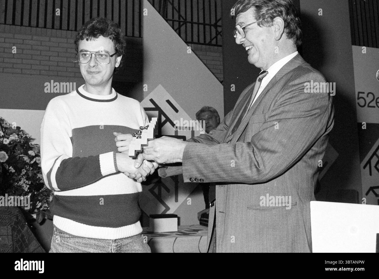 Verpflegung und Preisverteilung H.S.T. HST, Whizgle News, Dutch Desk, Niederlande, 1950 - 2000 am 28-01-1990. Dies sind die Elemente im Bild. In dieser Szene sind zwei Männer in einem Moment der Anerkennung und des Feierns verwickelt. Ein Mann auf der linken Seite trägt einen hellen Pullover mit kräftigen horizontalen Streifen in Rot und Dunkelblau, der einen auffälligen Kontrast zur formelleren Kleidung des Mannes neben ihm bildet. Er hat eine Brille und einen etwas zurückhaltenden Ausdruck, der leicht zur Seite steht, wenn er eine Auszeichnung oder Trophäe entgegennimmt. Der Mann rechts, gekleidet in Anzug und Krawatte, strahlt ein Gefühl des Autors aus Stockfoto