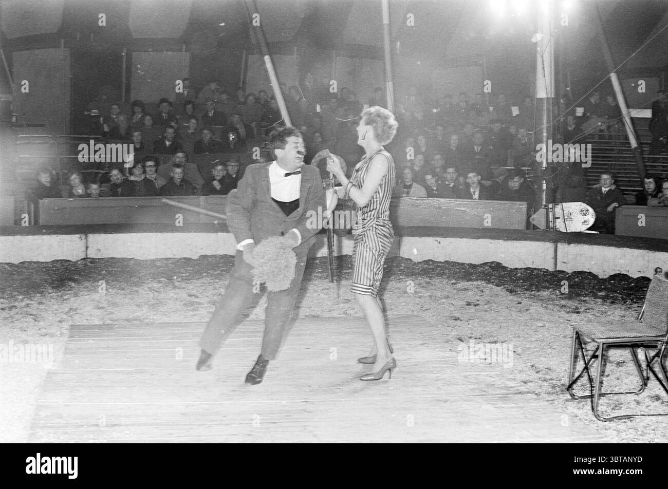 Toni Boltini Zirkusfilm Circus, Whizgle News, Dutch Desk, Niederlande, 1950 - 2000 am 10-04-1963. Die Abbildung zeigt diese Themen. Die Szene entfaltet sich in einer belebten Zirkusarena, voller Leben und Energie. In der Mitte hält ein Darsteller in einem klassischen Clown-Outfit – komplett mit übertriebenen Details und einem verspielten Auftreten – eine große, flauschige Requisite, die einen Hauch von Laune verleiht. Er wird mitten im Geschehen gefangen, vielleicht inmitten eines komischen Tanzes oder einer spielerischen Interaktion. Ihm gegenüber steht eine Frau, elegant gekleidet in einem auffälligen, glänzenden Kleid, das das Licht einfängt. Ihre Haltung und Expressi Stockfoto