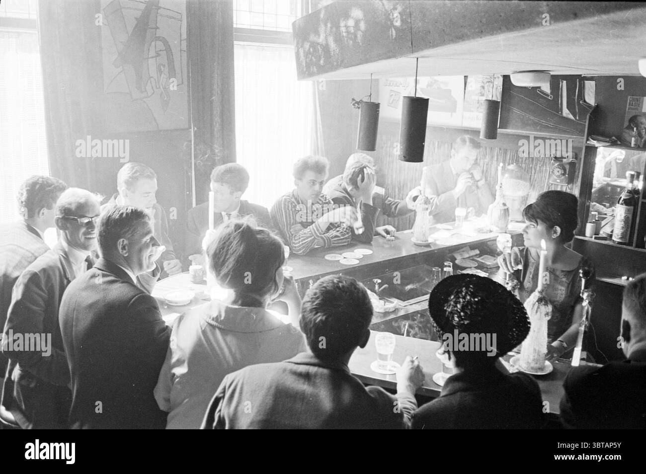 Pigalle Jansstraat Haarlem Cafe Haarlem Jansstraat the Netherlands, Whizgle News, Dutch Desk, Niederlande, 1950 - 2000 am 11-07-1963. Dies sind die Elemente im Bild. Die Szene fängt das lebendige Innere einer Bar ein, voller Menschen, die sich unterhalten und ihre Getränke genießen. Die Atmosphäre ist lebendig und dynamisch und spiegelt ein geselliges Treffen wider. Im Vordergrund befindet sich eine Gruppe elegant gekleideter Personen an der Bar, deren Gesichter von sanfter, diffuser Beleuchtung beleuchtet werden, die ein warmes Ambiente schafft. Die Männer tragen maßgeschneiderte Anzüge, einige mit Krawatten, während die Wo Stockfoto