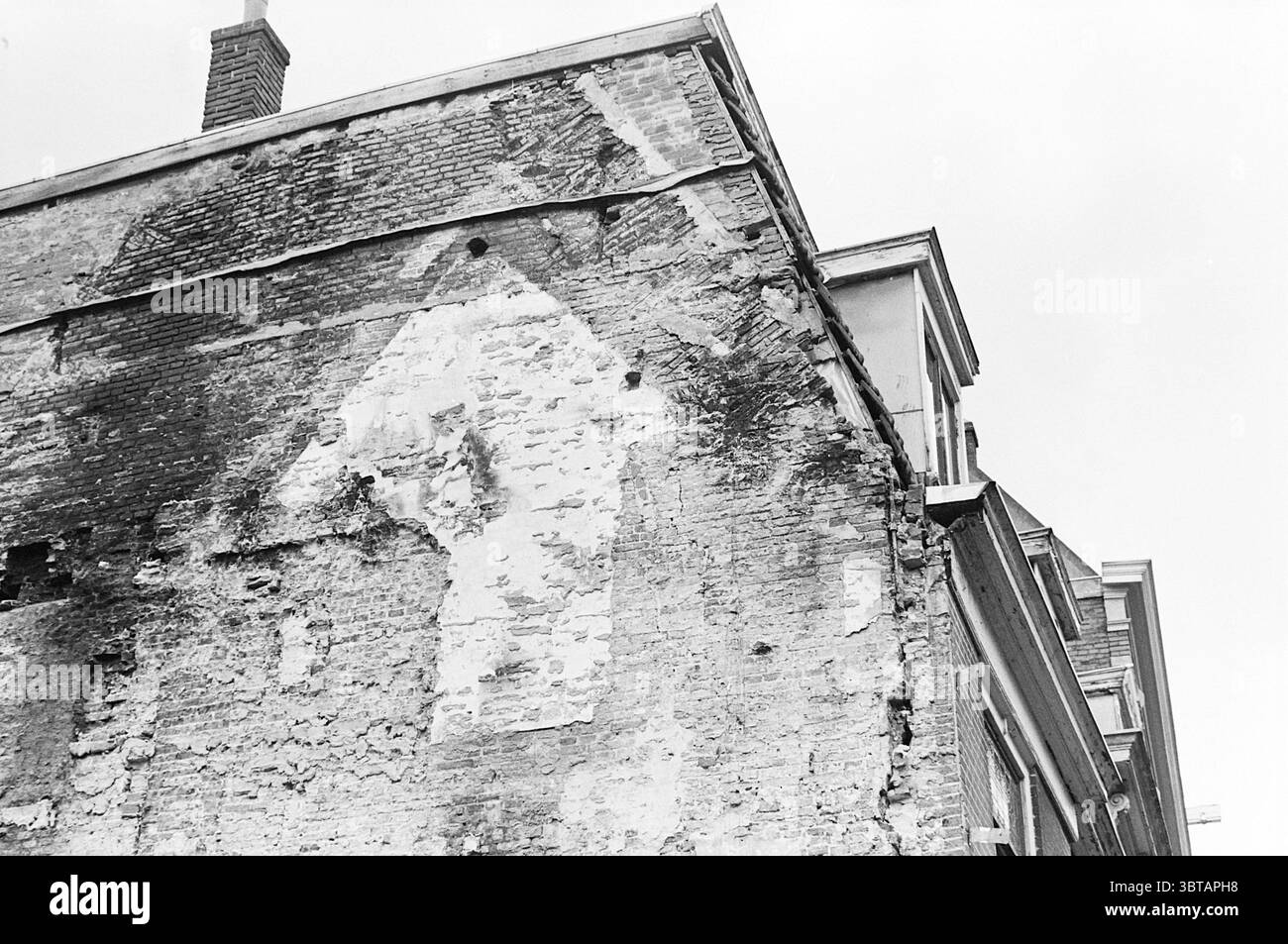 Risse in der Wand des Fa. Hensen Paarlaarsteeg Buildings Walls Haarlem Paarlaarsteeg Niederlande, Whizgle News, Dutch Desk, Niederlande, 1950 - 2000 am 19-11-1974. Das Bild enthält diese Themen. Die Szene zeigt eine verwitterte Wand eines Gebäudes, die sich durch ihre raue und sich verschlechternde Textur auszeichnet. Die Ziegelsteine sind in mehreren Bereichen sichtbar und zeigen eine Mischung aus Erdtönen, darunter tiefes Rot, Braun und Anklänge von verblasstem weiß, wo der Gips zerbröckelt ist. Die verblassenden Farben deuten auf Alter und Vernachlässigung hin und erwecken einen Sinn für Geschichte und Geschichten, die nicht erzählt werden. Über der Mauer befindet sich ein Teil des Gebäudes Stockfoto