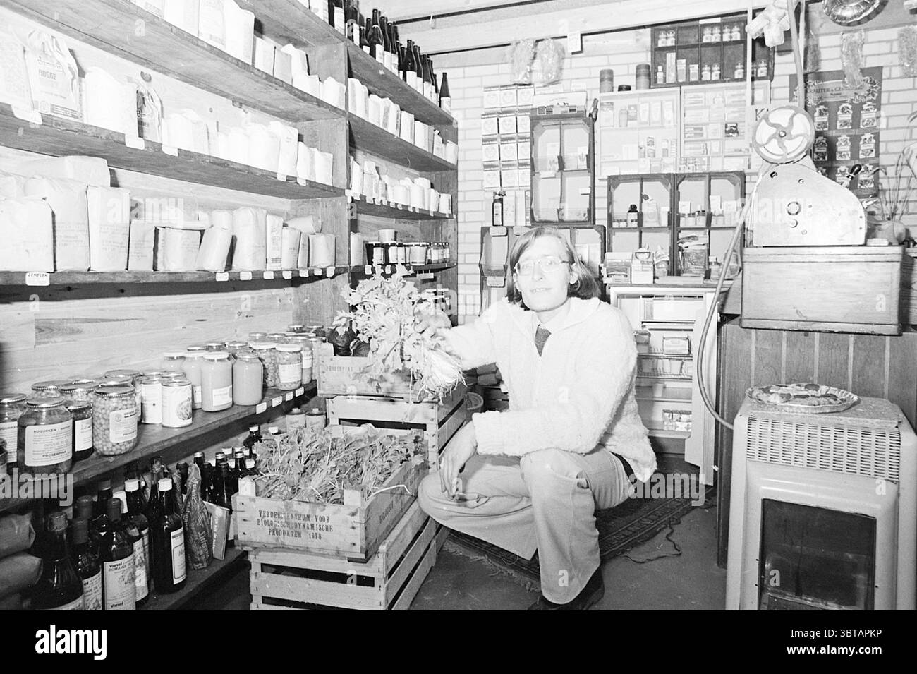 Int. Macro Bio Gemüsehändler Gemüse, Whizgle News, Dutch Desk, Niederlande, 1950 - 2000 am 17-04-1978. Dies sind die Elemente im Bild. Die Szene spielt in einem malerischen, rustikalen Geschäft, das ein Gefühl von Nostalgie weckt. Die Regale sind mit einer Vielzahl von Gläsern und Flaschen ausgestattet, deren Inhalt von bunten Konfitüren bis zu natürlichen Kräutern reicht und eine reiche Palette an Erdtönen bietet. Die Holzregale haben ein warmes, einladendes Finish, und Sie können eine Reihe von Vintage-Verpackungen sehen, die dem Raum Charakter verleihen. Im Vordergrund steht eine Person neben einer Holzkiste, die mit getrocknetem Plan gefüllt ist Stockfoto