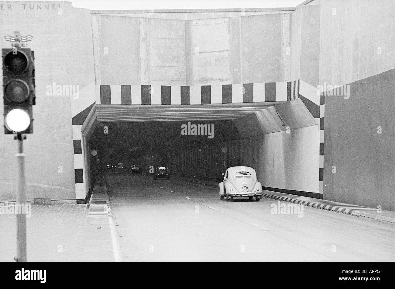 Marking North Entrance Velsertunnel Tunnels, Whizgle News, Dutch Desk, Niederlande, 1950 - 2000 am 12-02-1965. Diese Themen sind in der Abbildung dargestellt. Die Szene präsentiert eine krasse urbane Umgebung, die durch einen breiten Tunneleingang gekennzeichnet ist, der von auffälligen, kontrastierenden geometrischen Mustern in Schwarz und weiß eingerahmt wird. Das Gebäude hat ein industrielles Flair, mit Betonwänden, die sich über ihnen erheben und an einer sauberen, rechteckigen Öffnung aufeinander treffen. Im Inneren des Tunnels sind die Wände in glatten, gedämpften Tönen lackiert, wodurch ein Gefühl von Tiefe entsteht, das den Blick nach innen zieht. Die Beleuchtung ist schwach, was auf ein Gefühl von Einsamkeit hindeutet. Stockfoto