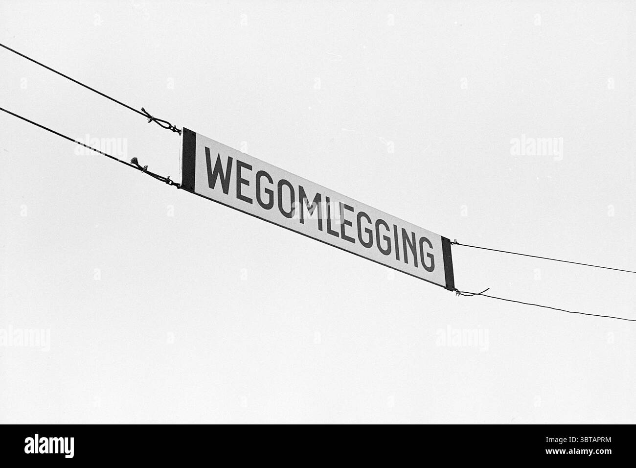 Collage Schiphol Schiphol, Whizgle News, Dutch Desk, Niederlande, 1950 - 2000 am 23-12-1970. Diese Themen werden in der Abbildung angezeigt. Die Szene zeigt eine schlichte, minimalistische Umgebung, die von einer kontrastreichen Schwarz-weiß-Ästhetik dominiert wird. Im oberen Teil hängt ein Schild an zwei dünnen Linien, das sich horizontal über den Rahmen erstreckt. Das Zeichen selbst wird gut sichtbar angezeigt, mit fett gedruckten Großbuchstaben und einer starken Schriftart, die seine Botschaft unterstreicht. Der Hintergrund ist mit einem weiten Himmel gefüllt, der durch einen weichen Verlauf gekennzeichnet ist, der von einem hellen weiß zu den Kanten übergeht. Stockfoto
