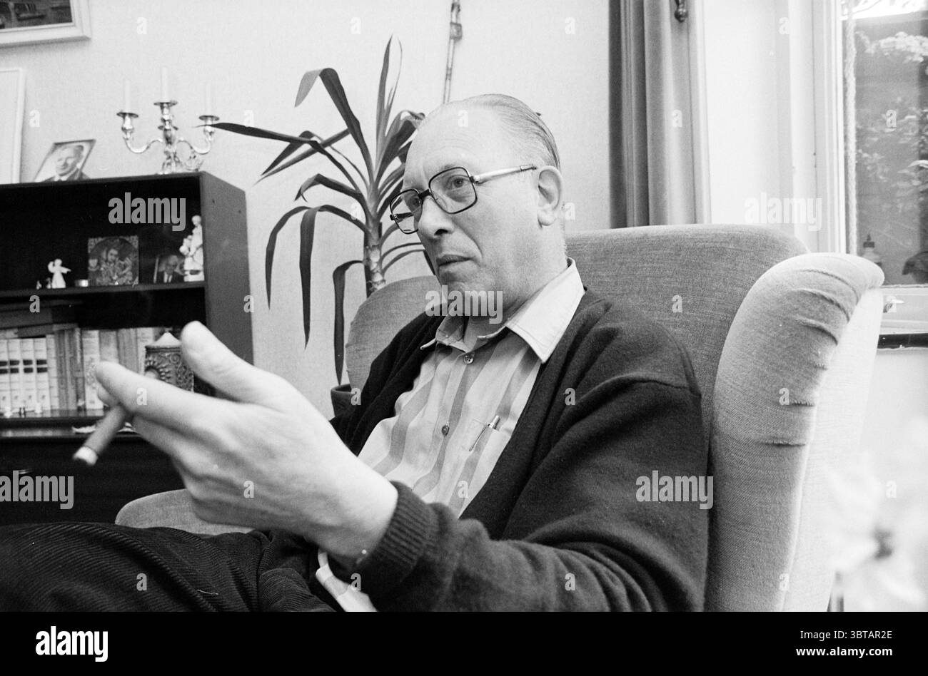 Roman Catholic Missionary v.d. Mountain Haarlem Waalstraat the Netherlands, Whizgle News, Dutch Desk, Niederlande, 1950 - 2000 am 09-06-1989. Das Bild enthält diese Themen. Die Szene spielt drinnen mit einem Mann mittleren Alters, der bequem in einem Sessel sitzt. Er trägt ein geknöpftes Hemd mit dezentem Streifenmuster, ergänzt durch eine dunkle Strickjacke, die einen gemütlichen Touch verleiht. Seine Brille gibt einen nachdenklichen Ausdruck, während er mit einer Hand Gesten macht, wahrscheinlich mitten im Gespräch. Der Hintergrund zeigt einen gut ausgestatteten Wohnbereich. Ein dunkler Holzschrank beherbergt verschiedene Objekte, die suggerieren Stockfoto