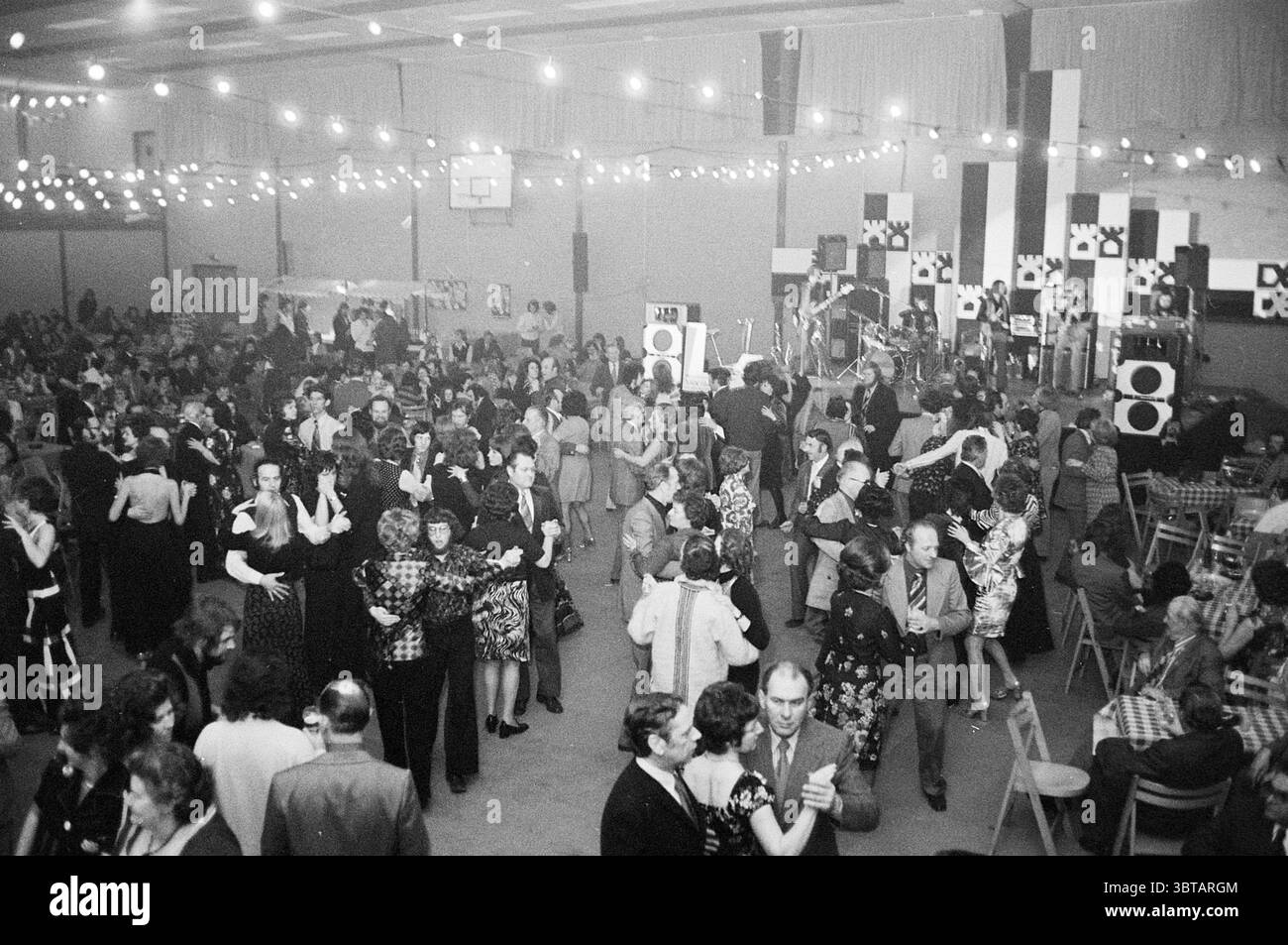 Farbenfroher Abend H.S.T.: Überblick über tanzende Menge + Mac und Katie Kissoon Blast Furnace Chess Tournament, Whizgle News, Dutch Desk, Niederlande, 1950 - 2000 auf 30-01-1975. Das Bild enthält diese Themen. Die Szene fängt ein lebhaftes Indoor-Event mit einer vielfältigen Begegnung von Menschen ein. Sie tragen eine Mischung aus formeller und halbformaler Kleidung, die auf einen festlichen Anlass, möglicherweise einen Tanz oder eine Feier, hinweist. Frauen in eleganten Kleidern wirbeln herum, während Männer in Anzügen und Krawatten sich in lebhaften Gesprächen und Tanzveranstaltungen unterhalten. Die Umgebung ist geräumig, mit einer hohen Decke und dekorierten Wänden, die Stockfoto