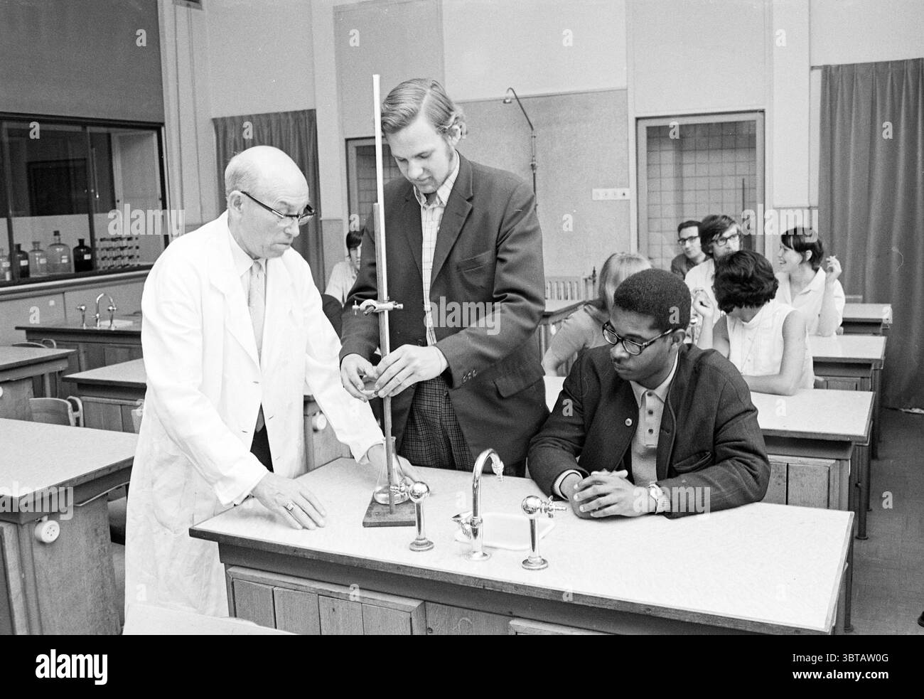 Reporting Analyst School Reporting Schools, Whizgle News, Dutch Desk, Niederlande, 1950 - 2000 am 24-06-1969. Dies sind die Themen im Bild. Die Szene spielt in einem Klassenzimmer, das für wissenschaftliche Experimente konzipiert ist. Die Umgebung ist geräumig, mit mehreren hölzernen Labortischen in Reihen angeordnet. Die Wände sind mit großen Fenstern verziert, die natürliches Licht hereinlassen und den Raum mit einem sanften Licht beleuchten. An vorderster Front sind zwei Männer an einer Demonstration beteiligt. Einer ist eine ältere Person, die einen weißen Labormantel trägt und Autorität und Erfahrung ausstrahlt. Er konzentriert sich intensiv auf ein Stück Labor-Equ Stockfoto