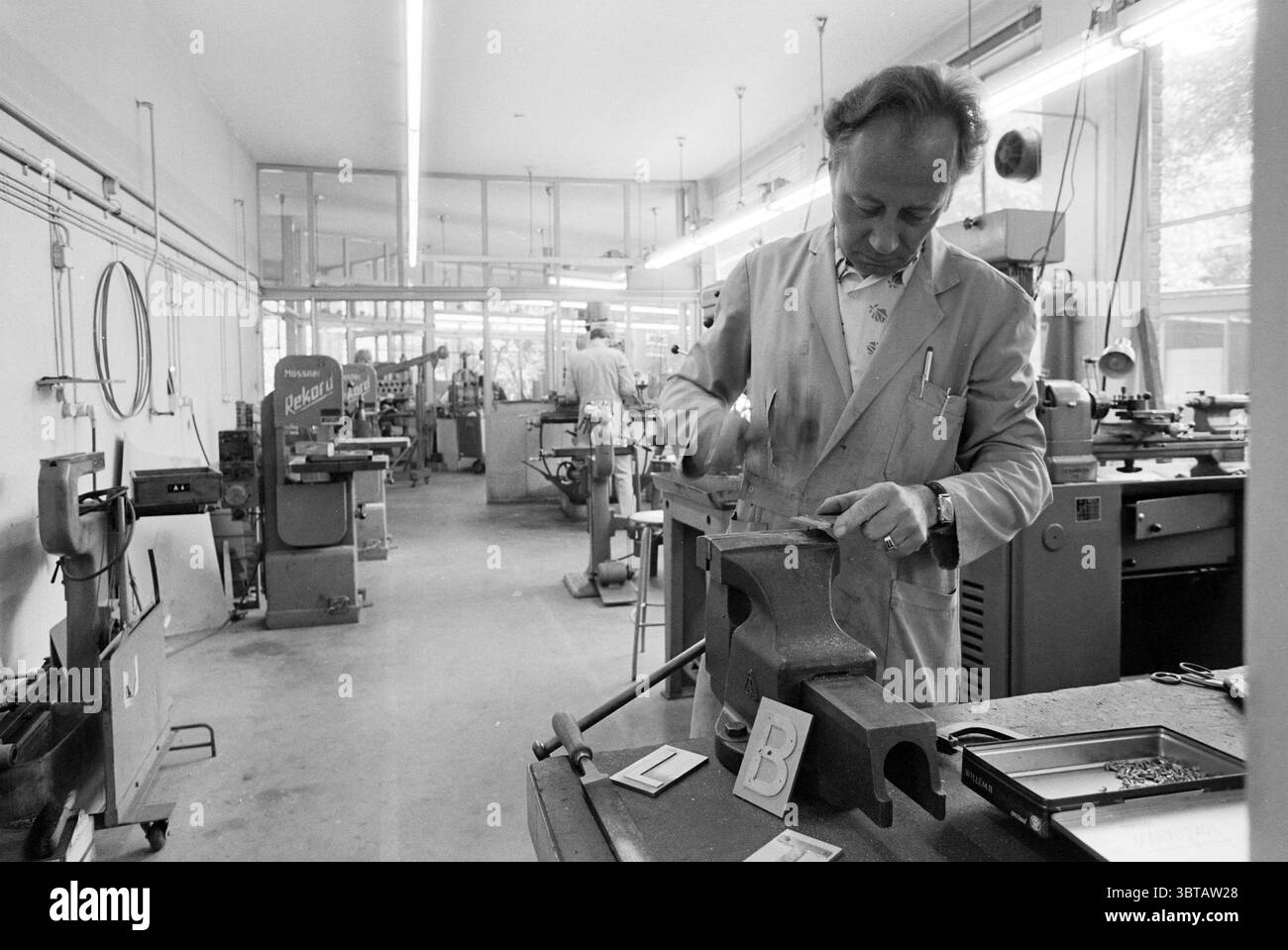 Metallverarbeitende Firma, Whizgle News, Dutch Desk, Niederlande, 1950 - 2000 am 06-1980. Dies sind die Themen im Bild. Die Szene zeigt eine geräumige Werkstatt mit verschiedenen Werkzeugen und Maschinen, die sich durch eine minimalistische und industrielle Ästhetik auszeichnet. Die vorherrschenden Farben sind gedämpfte Grau- und Weißtöne, die zu einer sauberen und funktionellen Atmosphäre beitragen. Im Vordergrund konzentriert sich ein Mann in einem hellen Laborkittel auf eine Aufgabe an einer Werkbank. Er benutzt einen Amboss, der seine Handwerkskunst zum Ausdruck bringt. Sein Ausdruck deutet auf Konzentration und Hingabe hin. Um ihn herum ist die Werkbank ein Stockfoto