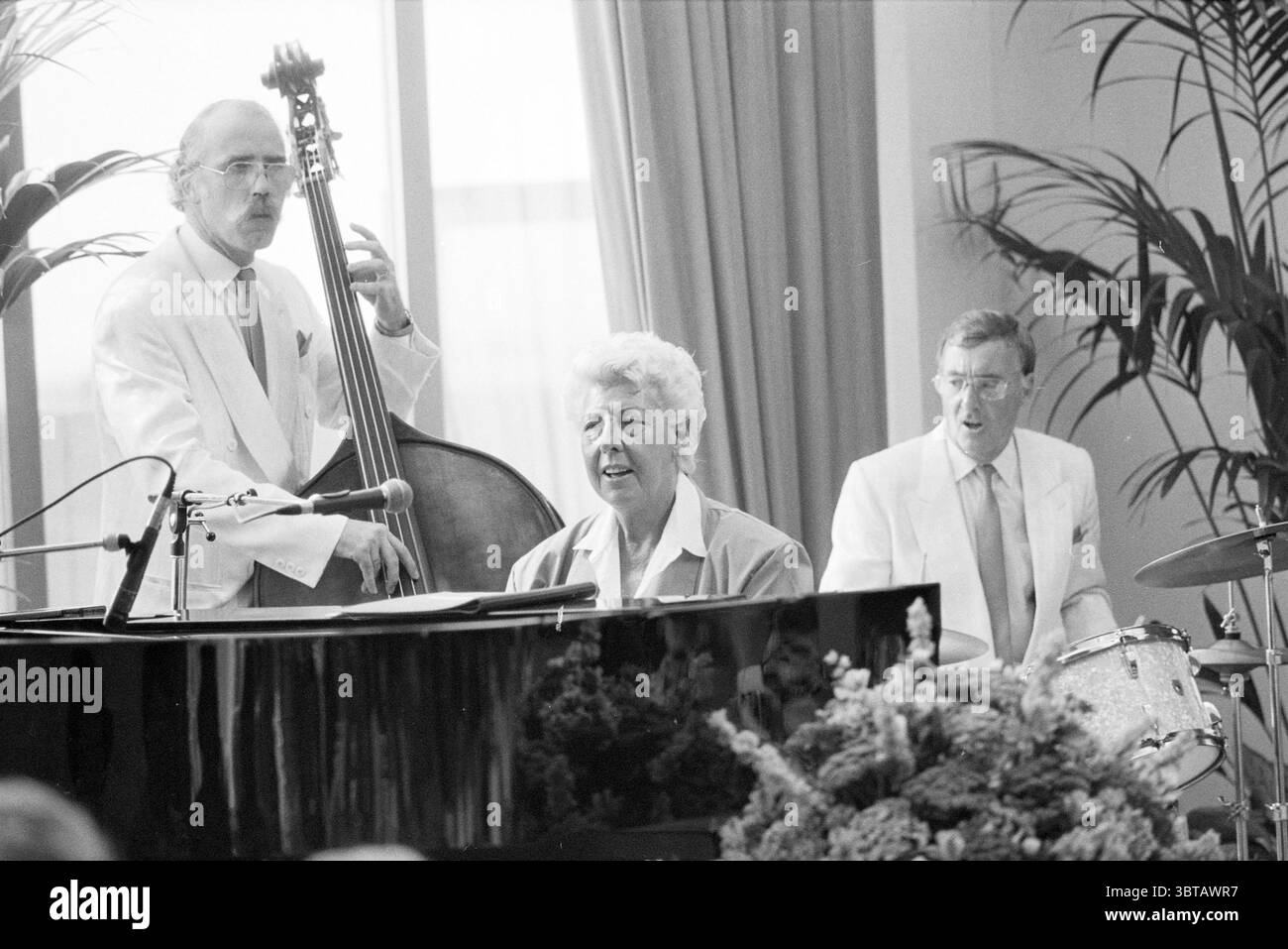 Strandhotel Pia Beck. Jazz, Whizgle News, Dutch Desk, Niederlande, 1950 - 2000 am 06-08-1989. Die Abbildung zeigt diese Themen. Die Szene bietet eine lebhafte musikalische Performance, bei der drei Musiker aktiv an ihrem Handwerk beteiligt sind. Die Einrichtung scheint ein elegant eingerichteter Innenraum zu sein, mit großen Fenstern, die weiches, diffuses natürliches Licht in den Raum einlassen und eine warme, aber gedämpfte Atmosphäre schaffen. Im Vordergrund glänzt ein Flügel mit einer polierten Oberfläche, die das Umgebungslicht und die umgebende Einrichtung reflektiert. Hinter dem Klavier sitzt eine Frau selbstbewusst am Keyboard, h Stockfoto