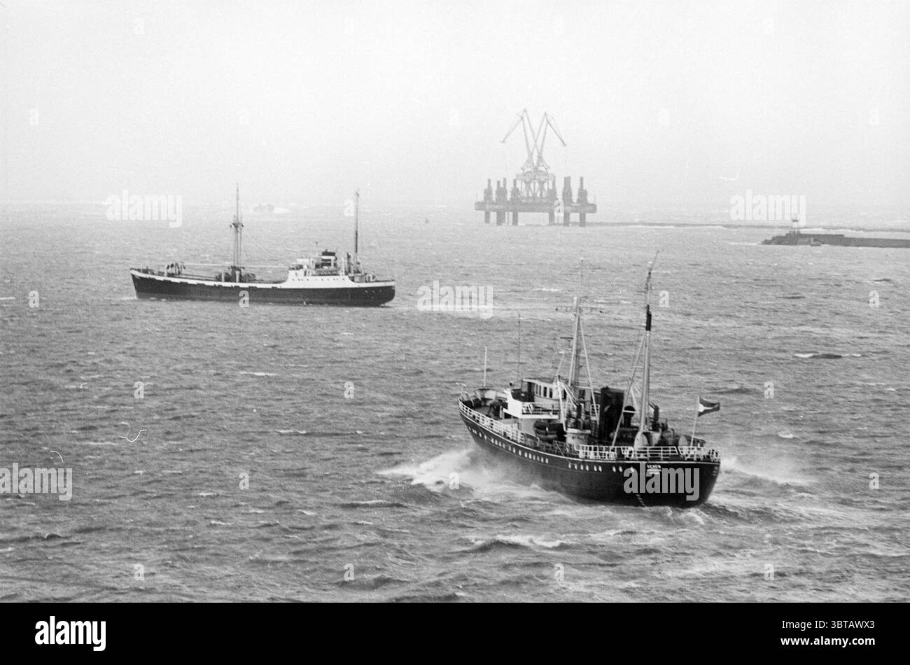 Zwei Schiffe verlassen den Hafen von IJmuiden., Whizgle News, Dutch Desk, Niederlande, 1950 - 2000. Dies sind die Elemente im Bild. Die Szene zeigt eine turbulente Meeresumgebung, die von bewölktem Himmel geprägt ist, der ein gedämpftes graues Licht über das Wasser wirft. Der Ozean wirkt abgehackt, wobei Wellen eine lebendige Oberflächenstruktur erzeugen, während sie gegeneinander wallen und aufprallen. Zwei Schiffe besetzen die Szene. Das erste, ein größeres Schiff, nimmt den Vordergrund ein und wird selbstbewusst durch die Wellen bewegt. Der Rumpf ist in dunklen Tönen gestrichen, die sich von den helleren Tönen des Meeres abheben. Das Schiff verfügt Stockfoto