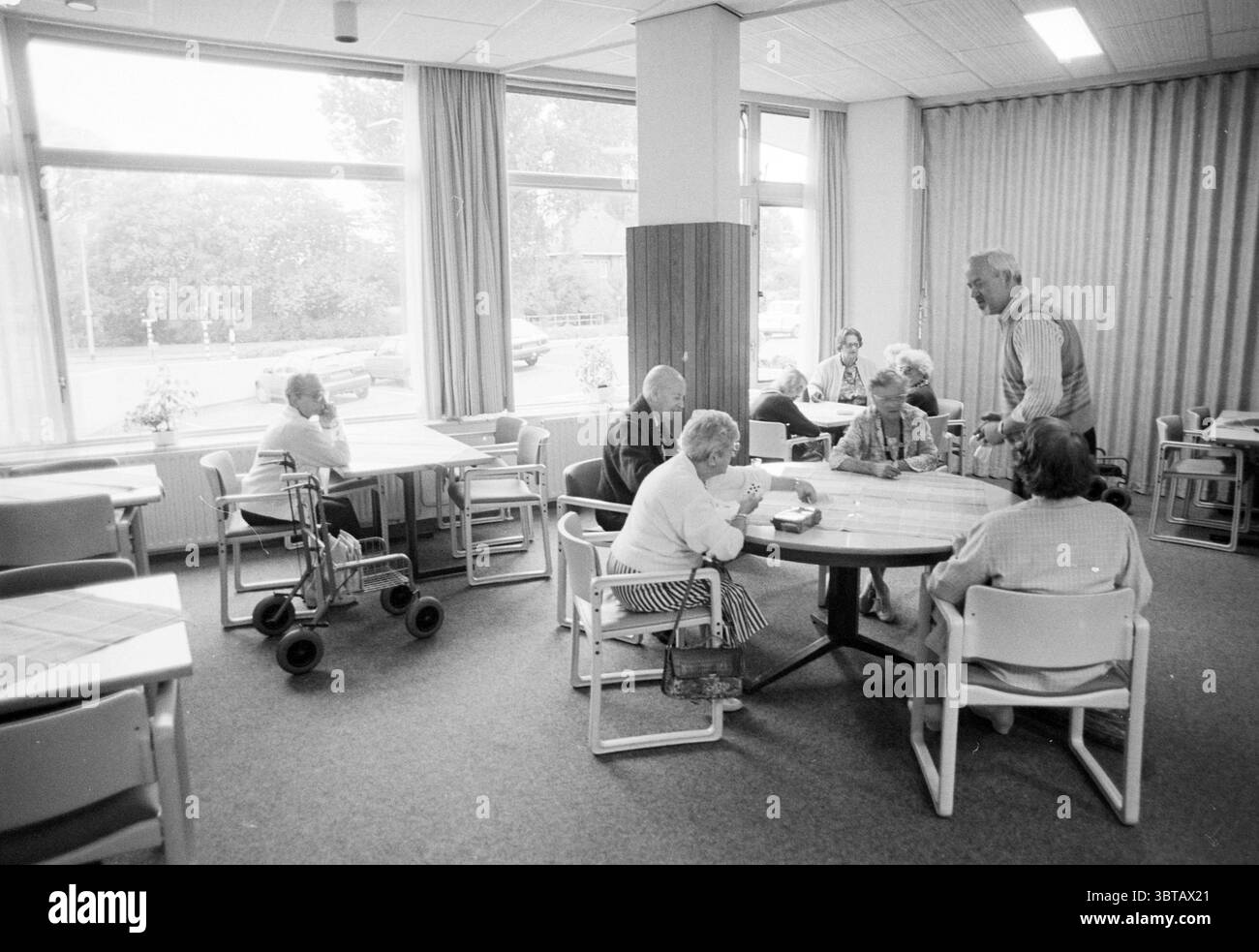 Konversationsraum Jacob in de Hout, Whizgle News, Dutch Desk, Niederlande, 1950 - 2000 am 17.09.1992. Das Bild enthält diese Themen. Die Szene zeigt einen Gemeinschaftsraum, wahrscheinlich innerhalb eines Pflegeheims oder einer Senioreneinrichtung, der durch helle Töne und eine luftige Atmosphäre gekennzeichnet ist. Mehrere Tische sind geräumig angeordnet, sodass sie leicht bewegt werden können. An vorderster Front führt eine Gruppe von sechs älteren Personen Gespräche an einem runden Tisch. Sie zeigen unterschiedliche Ausdrucksformen – einige hören aufmerksam zu, andere scheinen einen unbeschwerten Moment zu teilen. Eine Person steht ab Stockfoto