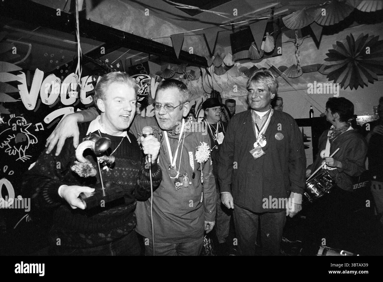 Festival der Karnevalslieder, Whizgle News, Dutch Desk, Niederlande, 1950 - 2000 am 02-02-1993. Die Abbildung zeigt diese Themen. Die Szene fängt eine lebhafte Feier ein, geprägt von einer dynamischen Atmosphäre voller Freude und Begeisterung. Eine Gruppe von drei Personen steht im Vordergrund. Die zentrale Figur in einem dunklen Pullover hält eine Trophäe hoch, sein Ausdruck ist lebhaft vor Aufregung und einem Hauch von Stolz. Er ist eindeutig der Mittelpunkt, der mit einem Lächeln Aufmerksamkeit erregt, das Energie ausstrahlt. Zu seiner Rechten steht ein Mann mittleren Alters, der eine Brille trägt und ebenfalls vor Freude strahlt. H Stockfoto