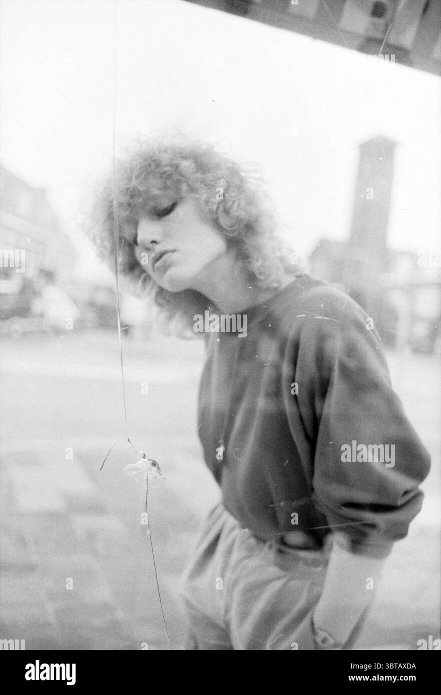 Einschubloch im Schaufenster Buntglas Blasglas im Allgemeinen, Whizgle News, Dutch Desk, Niederlande, 1950 - 2000 am 28-12-1983. Dies sind die Themen im Bild. Die Szene fängt ein junges Individuum ein, das in einer leicht verschwommenen urbanen Umgebung steht. Ihr Gesicht wird von weichen, voluminösen Locken eingerahmt, die ihrem Aussehen eine verspielte Qualität verleihen. Ihr Ausdruck ist kontemplativ und leicht geneigt, wodurch eine intime Verbindung zum Betrachter entsteht. Die Person trägt einen locker sitzenden, dunklen Pullover, der sich mit den helleren Farben ihrer Umgebung abhebt. Die Umgebung scheint zu sein Stockfoto