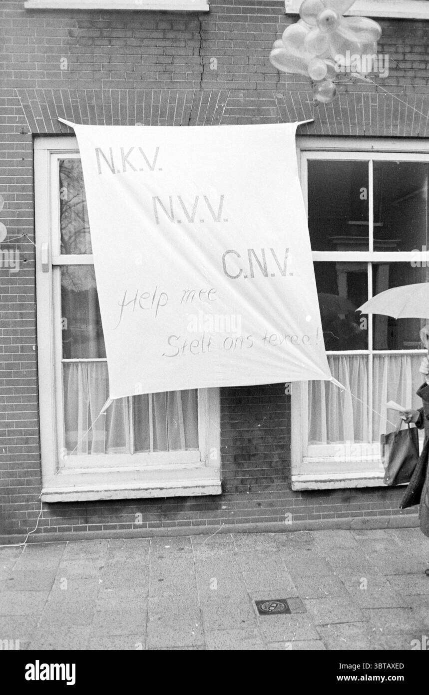 Banner Calling for the union., Whizgle News, Dutch Desk, Niederlande, 1950 - 2000. Das Bild enthält diese Themen. Die Umgebung scheint eine urbane Straßenszene zu sein, die durch eine Ziegelfassade aus Rot- und Brauntönen gekennzeichnet ist. Das Gebäude verfügt über große Fenster mit weißen Rahmen, was auf eine bescheidene, aber dennoch zugängliche Architektur hindeutet. Vor dem Gebäude hängt ein großes weißes Banner, das leicht weht, als ob es von einer sanften Brise aufgerührt würde. Die Oberfläche des Banners ist glatt und bildet einen starken Kontrast zum Backsteinhintergrund. Die allgemeine Stimmung, die durch das Banner und seine Botschaft vermittelt wird Stockfoto