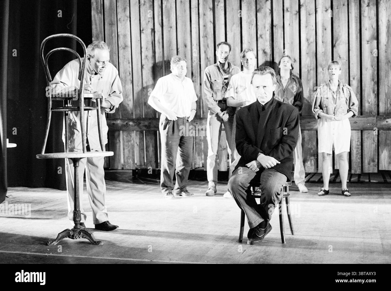 Probe 'Haarlem Theater' Haarlem the Netherlands, Whizgle News, Dutch Desk, Niederlande, 1950 - 2000 am 22-08-1991. Dies sind die Themen im Bild. Die Szene entfaltet sich in einer rustikalen, hölzernen Umgebung, die von einer Kulisse aus vertikalen Holzdielen geprägt ist, die ein Gefühl von Wärme und Alter ausstrahlen. Ein weiches, gedämpftes Licht fließt auf die Bühne und wirft sanfte Schatten, die die Texturen des Holzes und der Figuren verbessern. Im Vordergrund sitzt ein Mann auf einem einfachen, dunklen Holzstuhl, sein Ausdruck besinnlich, was auf ein Gefühl von Verletzlichkeit oder Introspektion hindeutet. Er ist im Dunkeln gekleidet Stockfoto