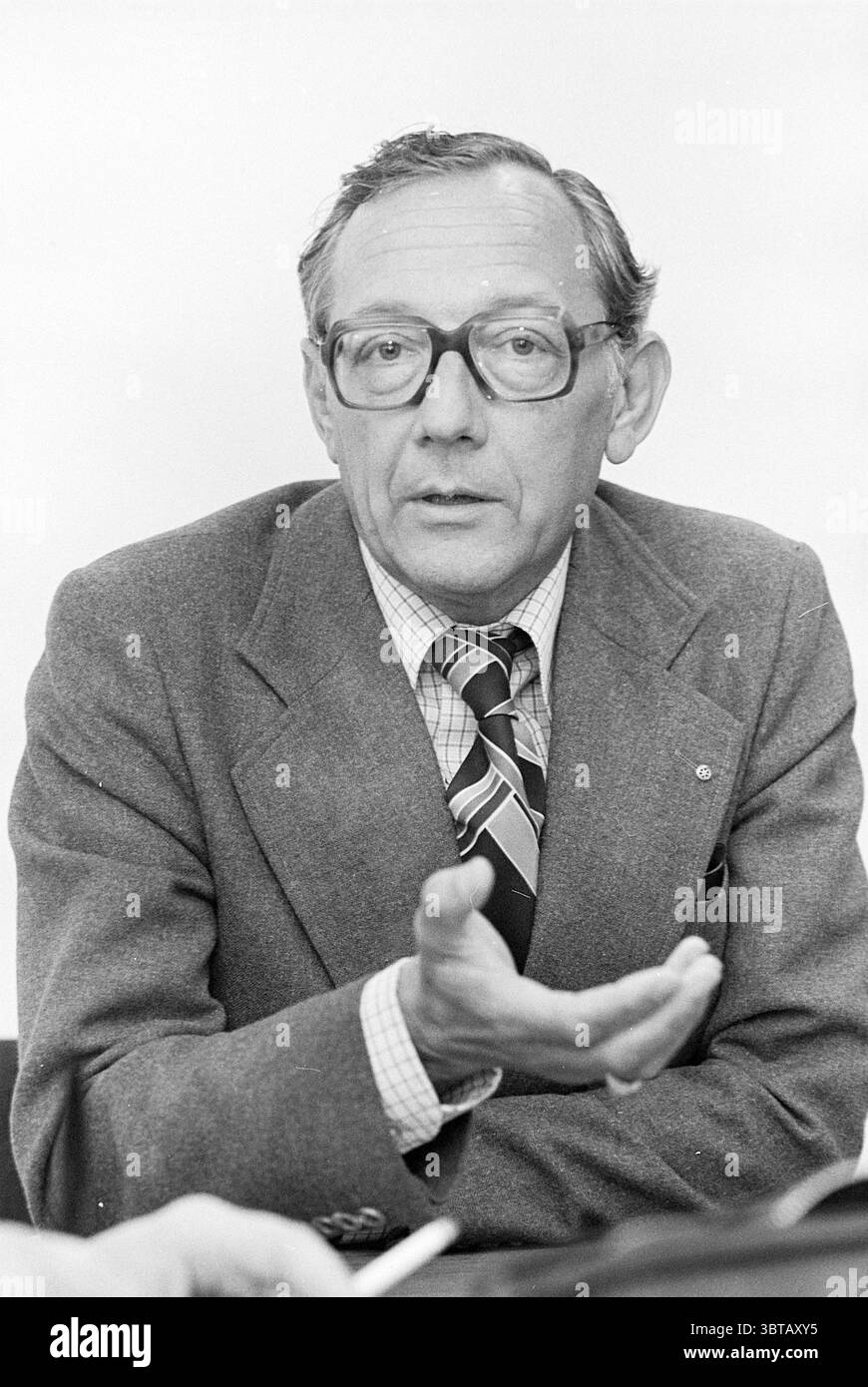 Porträt Mr. Kruitwagen Heemstede Porträts, Whizgle News, Dutch Desk, Niederlande, 1950 - 2000 am 19-08-1977. Das Bild enthält diese Themen. In der Szene sitzt ein Mann mittleren Alters an einem Tisch und führt ein Gespräch. Er trägt einen formellen, grauen Anzug, der durch eine gestreifte Krawatte ergänzt wird, die eine reiche Farbpalette aufweist, darunter tiefes Rot und Blau. Der Anzug wirkt maßgeschneidert und vermittelt einen professionellen Eindruck. Sein Hemd ist hell in der Farbe und bietet einen schönen Kontrast zum dunkleren Anzug. Der Ausdruck des Mannes vermittelt Aufmerksamkeit und Ernsthaftigkeit, während er mit einer Hand Gesten, Emph Stockfoto