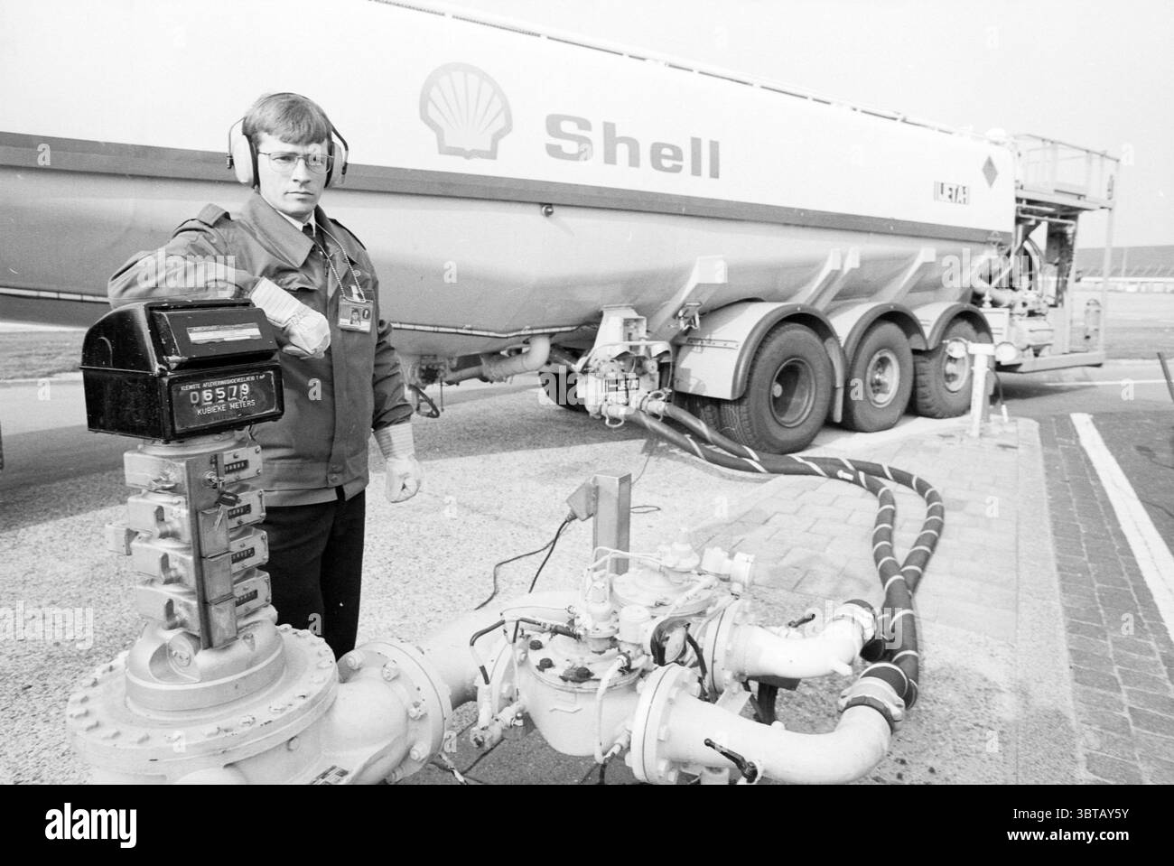 Schiphol Work Shell Tanker Schiphol, Whizgle News, Dutch Desk, Niederlande, 1950 - 2000 am 25-04-1991. Dies sind die Elemente im Bild. Die Szene zeigt einen Mann, der neben einem großen Tankwagen steht, der in erster Linie weiß ist und mit einem markanten Logo in leuchtendem Rot und Gelb verziert ist. Die starke Präsenz des Staplers dominiert den Hintergrund und deutet auf industrielle Funktionalität hin. Die Person trägt ein praktisches Outfit, das sich durch eine dunkle Jacke auszeichnet, die eine Arbeitsuniform andeutet, sowie Kopfhörer, die Kommunikations- oder Sicherheitsmaßnahmen anzeigen. Sein Ausdruck erscheint seri Stockfoto