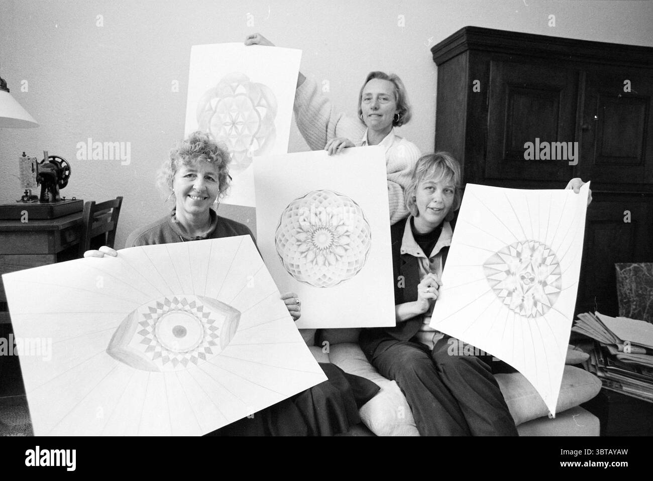 Kursleiter Chakra Zeichnungen Heemstede Kurse Zeichnungen Heemstede Niederlande, Whizgle News, Dutch Desk, Niederlande, 1950 - 2000 am 10-03-1989. Dies sind die Themen im Bild. In einem gemütlichen Innenraum versammeln sich drei Frauen, von denen jede große, aufwendig gestaltete Papierkunstwerke hält. Die Umgebung strahlt eine warme, einladende Atmosphäre aus, mit sanfter, diffuser Beleuchtung, die die Farben ihrer Kunstwerke unterstreicht. Die Wände sind hell und bieten eine dezente Kulisse, die die farbenfrohen Designs hervorhebt. Die erste Frau sitzt im Vordergrund und lächelt begeistert Stockfoto