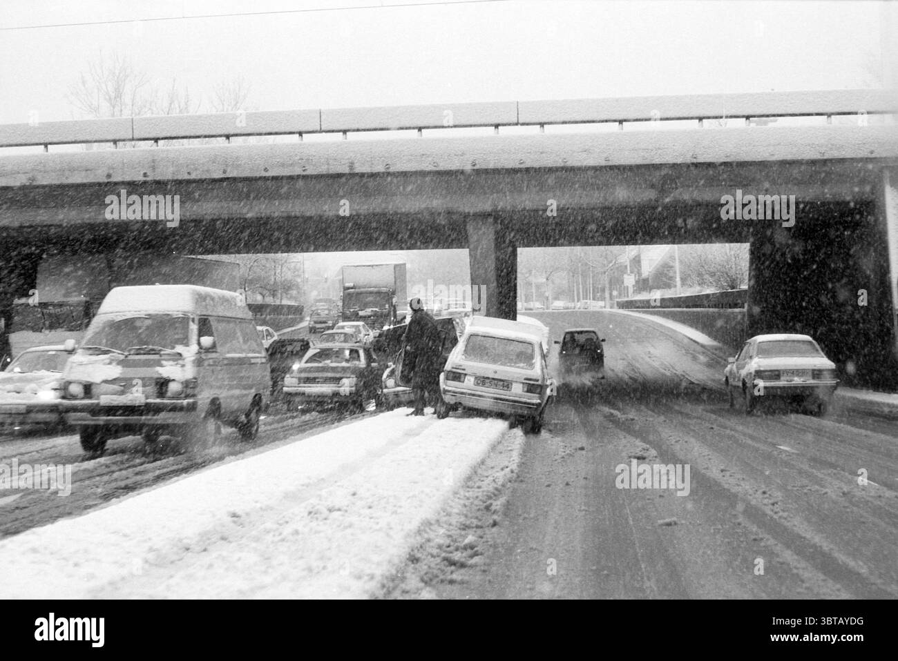 Autounfall im Schnee auf der Autobahn., Whizgle News, Dutch Desk, Niederlande, 1950 - 2000. Das Bild enthält diese Themen. Die Szene zeigt eine winterliche Stadtlandschaft unter starkem Schneefall. Eine Autobahn oder eine Überführung dominiert den Hintergrund, unter der Fahrzeuge in einem offensichtlichen Stau geraten. Die Straße ist mit einer dicken Schneedecke bedeckt, und einige Abschnitte erscheinen tückisch eisig. Die Fahrzeuge sind über die gesamte Komposition verteilt, mit einer Mischung aus Farben wie weiß, Grau und dunkleren Schattierungen, die einen Kontrast zum hellweißen Schnee bilden. Einige Autos haben Schnee angesammelt Stockfoto Autounfall im Schnee auf der Autobahn., Whizgle News, Dutch Desk, Niederlande, 1950 - 2000. Das Bild enthält diese Themen. Die Szene zeigt eine winterliche Stadtlandschaft unter starkem Schneefall. Eine Autobahn oder eine Überführung dominiert den Hintergrund, unter der Fahrzeuge in einem offensichtlichen Stau geraten. Die Straße ist mit einer dicken Schneedecke bedeckt, und einige Abschnitte erscheinen tückisch eisig. Die Fahrzeuge sind über die gesamte Komposition verteilt, mit einer Mischung aus Farben wie weiß, Grau und dunkleren Schattierungen, die einen Kontrast zum hellweißen Schnee bilden. Einige Autos haben Schnee angesammelt Stockfoto