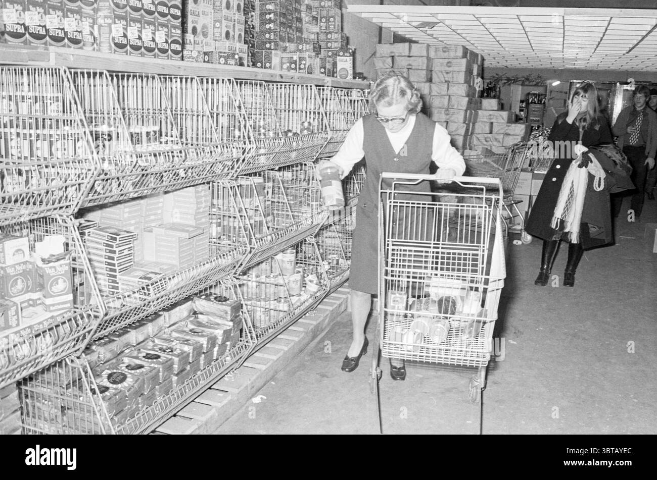 Preisverleihung Kongro Award Ceremony, Whizgle News, Dutch Desk, Niederlande, 1950 - 2000 am 30-12-1971. Diese Themen werden in der Abbildung angezeigt. In einem belebten Lebensmittelgeschäft fängt die Szene eine Frau ein, die mit ihrer Einkaufsroutine beschäftigt ist. Sie hat schulterlange Haare, die praktisch gestylt sind, und trägt ein dunkles Kleid mit weißem Kragen, was ihr einen klassischen, zeitlosen Look verleiht. Sie kniet neben einem Einkaufswagen und untersucht sorgfältig eine Dose, ihre Konzentration ist offensichtlich. Der Einkaufswagen ist teilweise mit verschiedenen Lebensmitteln gefüllt, und die Regale neben ihr sind mit einer Reihe von verpackten Güllen ausgekleidet Stockfoto