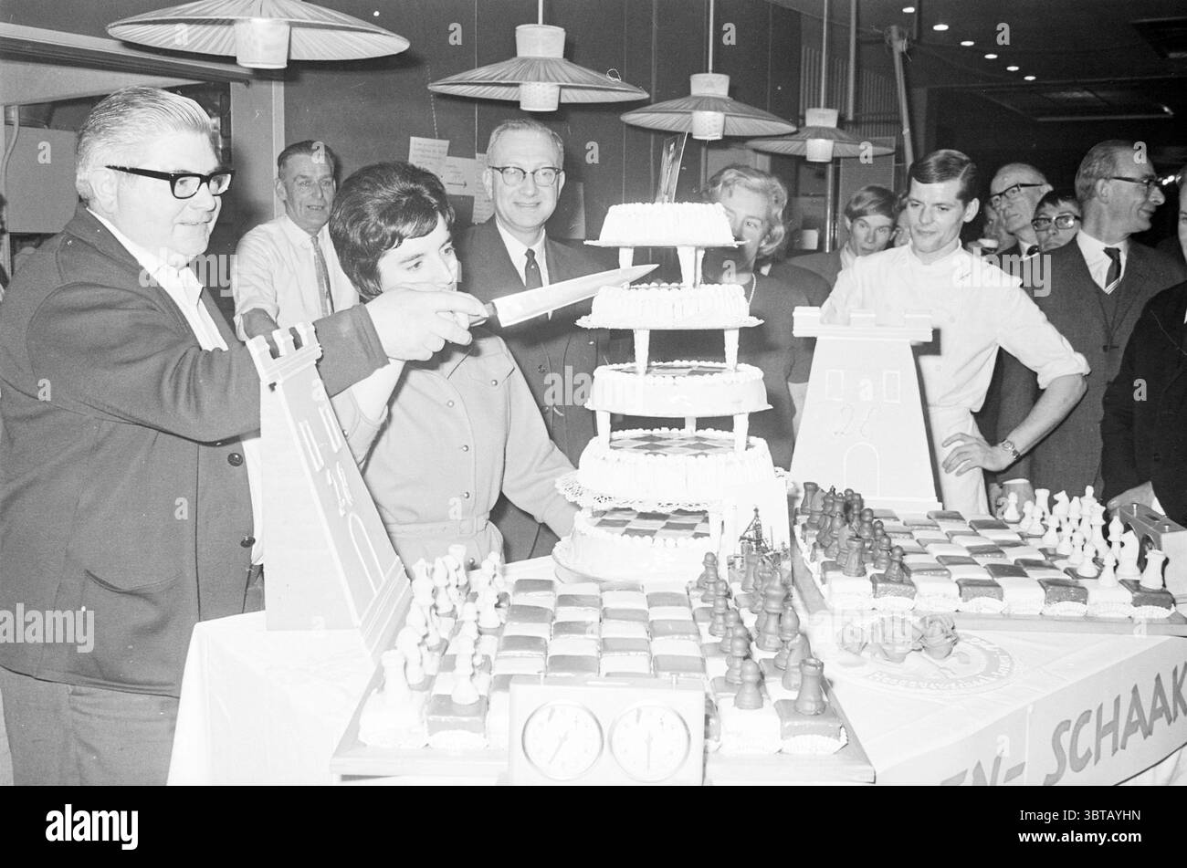 Blast Furnace Chess Tournament: Frau schneidet eine „Hochzeitstorte“, begleitet von zwei Schokoladenschachbrettern, Whizgle News, Dutch Desk, Niederlande, 1950 - 2000 am 01-1967. Dies sind die Themen im Bild. Die Szene ist lebhaft und festlich, um einen großen, mehrstufigen Kuchen, der so dekoriert ist, dass er ein Schachthema widerspiegelt. Die Schichten des Kuchens sind weiß mit vielleicht einigen komplizierten Mustern, die auf eine Feier mit Schach hinweisen, was durch die Verwendung von Schachstücken und einem Schachbrett aus Konfekt angezeigt wird. Um den Kuchen herum sind mehrere Leute, einschließlich eines gut gekleideten Mannes mit Brille, der ist Stockfoto
