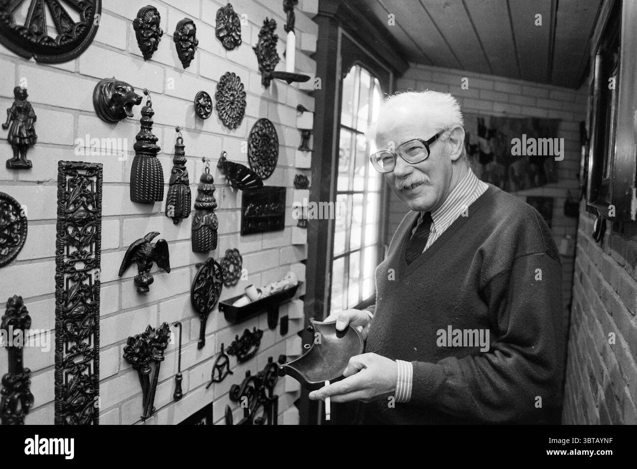 Architekt van den Akker Zwanenburg, Whizgle News, Dutch Desk, Niederlande, 1950 - 2000 am 20-02-1991. Diese Themen werden in der Abbildung angezeigt. Die Szene zeigt einen älteren Herrn in einem warmen, einladenden Raum mit einer Vielzahl dekorativer Objekte. Er steht in der Nähe einer Wand, die mit einer eklektischen Sammlung von Metallornamenten und Skulpturen geschmückt ist, die komplizierte Details und unterschiedliche Texturen zeigen und einen visuell reichen Kunstschmuck schaffen. Die Objekte umfassen verschiedene Formen und Figuren, von Vögeln über Blumen bis hin zu abstrakten Designs, die alle subtile Schatten auf die helle Ziegelwand werfen. Stockfoto