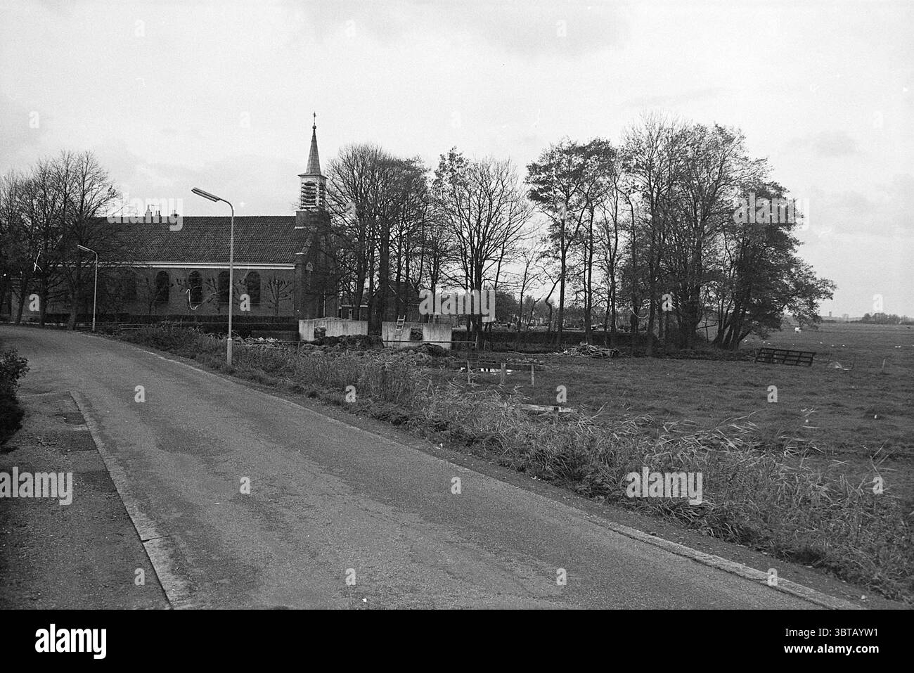 Kirche (außen)., Whizgle News, Dutch Desk, Niederlande, 1950 - 2000. Diese Themen werden in der Abbildung angezeigt. Die Szene präsentiert eine ruhige ländliche Landschaft unter einem bewölkten Himmel, die eine ruhige und doch düstere Stimmung hervorruft. Im Vordergrund zieht sich eine kurvige Straße diagonal, die den Blick des Betrachters in den Hintergrund führt. Der Asphalt ist leicht abgenutzt und weist Risse und Flecken auf, die auf eine jahrelange Nutzung hindeuten. Auf der einen Seite der Straße steht eine kleine Kirche. Sein Turm mit klassischem spitzem Design reicht nach oben und ergänzt die vertikalen Linien, die von umliegenden Bäumen geschaffen werden. Die Kirche ist AR Stockfoto