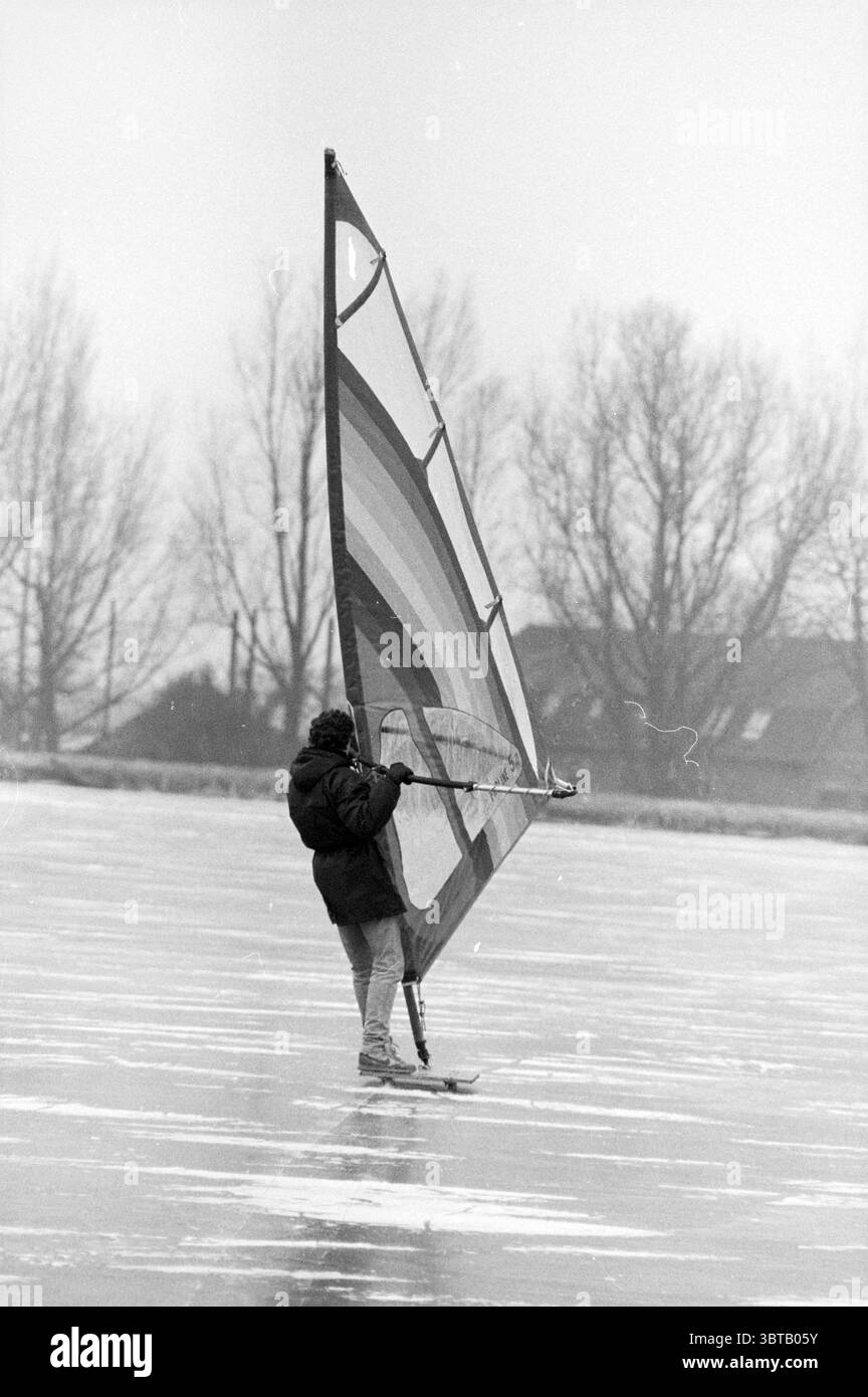 Eissurfen., Whizgle News, Dutch Desk, Niederlande, 1950 - 2000. Das Bild enthält diese Themen. Die Szene zeigt eine Einzelfigur, die auf einer riesigen, gefrorenen Weite steht. Die Person trägt eine dunkle, sperrige Jacke mit Kapuze, die einen starken Kontrast zur vereisten Oberfläche bildet. Ihre Haltung ist dynamisch und deutet auf Bewegung hin, während sie an einem großen, bunten Segel festhalten, das mit kräftigen horizontalen Streifen verziert ist, die leuchtende Rot-, Gelb-, Grün- und Blautöne zeigen. Der Hintergrund ist minimal mit blattlosen Bäumen bestückt, deren Äste sich in Skelettform nach außen erstrecken, was auf Ea hindeutet Stockfoto