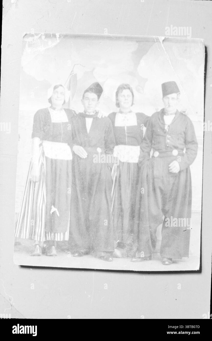 Menschen in Volendam Kostüm., Whizgle News, Dutch Desk, Niederlande, 1950 - 2000. Diese Themen werden in der Abbildung angezeigt. In der Szene stehen vier Personen zusammen, eng und freundlich angeordnet, was auf eine gemeinsame Verbindung hinweist. Sie sind in traditioneller Kleidung gekleidet, die ein bestimmtes kulturelles Erbe widerspiegelt. Die erste Person auf der linken Seite trägt einen langen Rock mit vertikalen Streifen, dazu eine weiße Bluse und ein Kopftuch, das einen Hauch von Eleganz verleiht. Die zweite Person steht neben ihr in einem schwarzen Kleid mit einem taillierten Oberteil und einer auffälligen Schleife, was auf eine andeutet Stockfoto