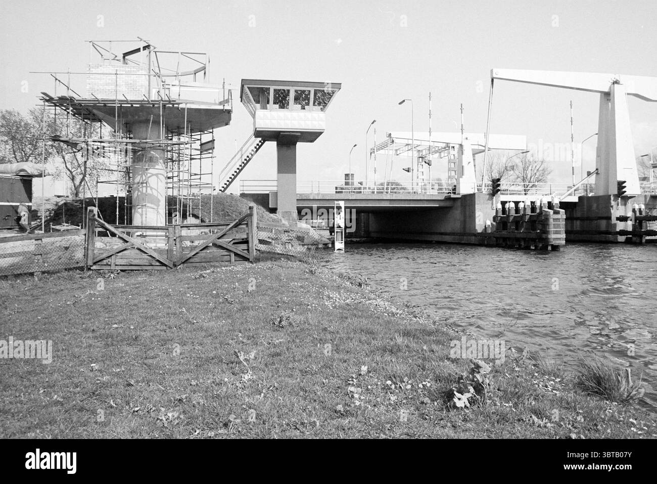 Neues Brückenhaus Cruquius Cruquius, Whizgle News, Dutch Desk, Niederlande, 1950 - 2000 am 26-04-1992. Das Bild enthält diese Themen. Die Szene zeigt eine Kanalkulisse, in der eine Brücke das Wasser unter sich spannt. Auf der einen Seite führt ein grasbewachsenes Gebiet mit spärlichen Flecken von Wildblumen zum Wasserrand, was einen Hinweis auf die Präsenz der Natur in der bebauten Umgebung gibt. Links erhebt sich ein teilweise gebautes Bauwerk, das von Gerüsten ummantelt ist. Das Gebäude wirkt modern und funktional und beherbergt möglicherweise eine Art Betriebskontrolle, die durch seine schlanken, geometrischen Linien hervorgehoben wird. Das Substrat von Stockfoto