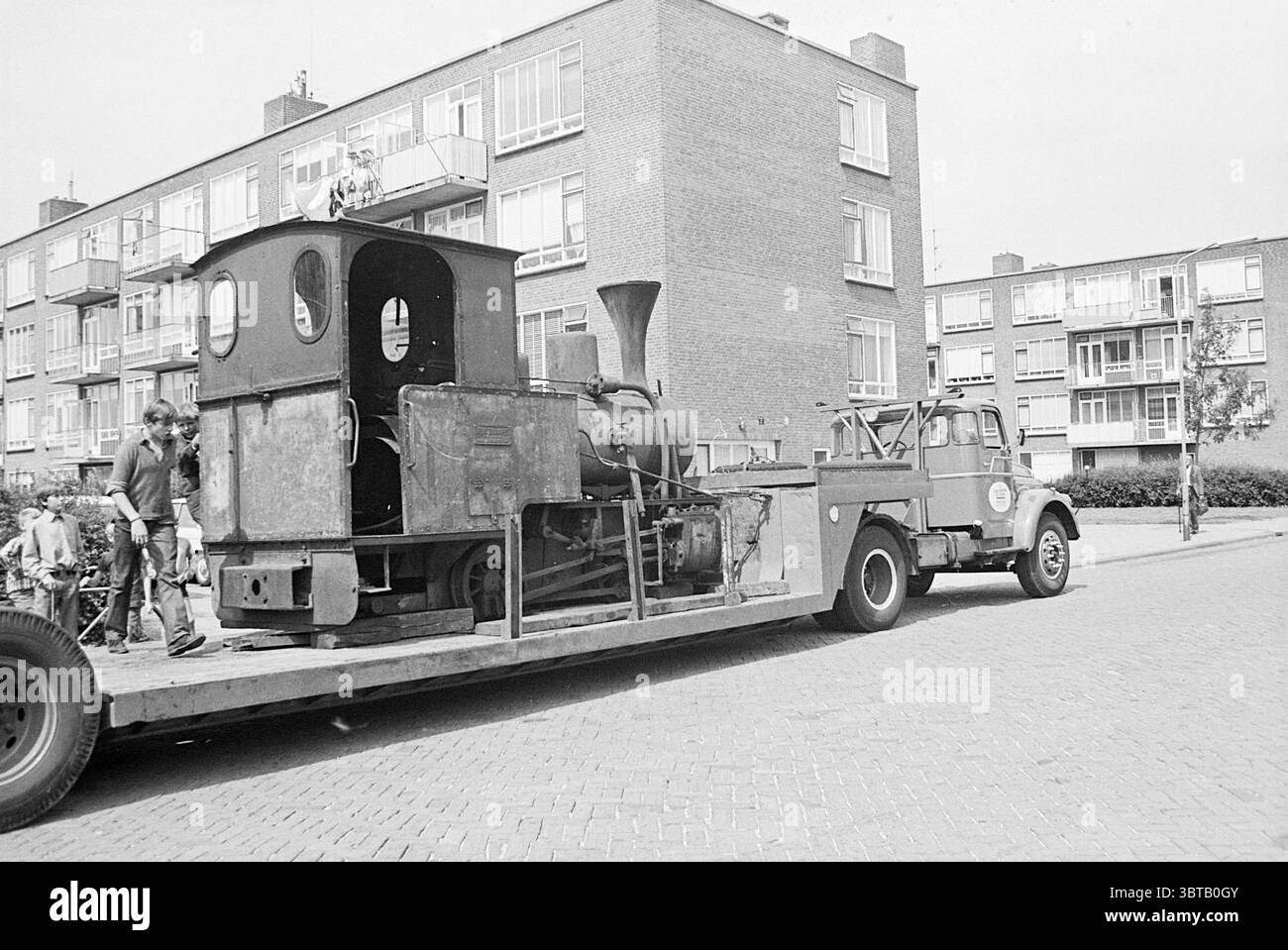Abfahrt Lokomotive Departure, Whizgle News, Dutch Desk, Niederlande, 1950 - 2000 am 07-08-1967. Dies sind die Elemente im Bild. Die Szene zeigt eine klassische Dampfmaschine auf einem Tieflader, die ihr industrielles Design mit einem markanten Schornstein und verwittertem Äußeren zeigt. Der Zug scheint bewusst transportiert worden zu sein, was auf ein Gefühl von Nostalgie oder historischer Bedeutung hinweist. Um dieses zentrale Objekt herum erheben sich Wohngebäude im Hintergrund, die sich durch ihr blockiges, zweckmäßiges Design und eine gedämpfte Farbpalette aus hellen Grau- und Brauntönen auszeichnen. Server Stockfoto