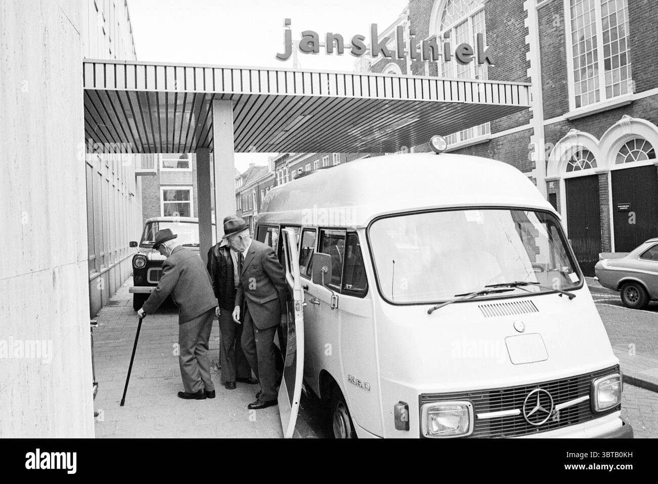 Ankunft von Patienten bei Janskliniek Zieken en Ziekenhuizen, Whizgle News, Dutch Desk, Niederlande, 1950 - 2000 am 30-06-1977. Die Abbildung zeigt diese Themen. Die Szene zeigt eine geschäftige urbane Umgebung, die vor allem durch eine große, geometrische Gebäudefassade geprägt ist, die ein klares, modernistisches Design hat. Das Äußere ist überwiegend hellgrau mit einer strukturierten Oberfläche, die eine subtile Tiefe verleiht. An der Vorderseite führt ein breiter Betonweg zum Eingang, der etwas von einem überhängenden Dach beschattet wird. Im Vordergrund steht ein weißer Van, vielleicht ein medizinisches Transportfahrzeug, in einem Winkel mit Stockfoto