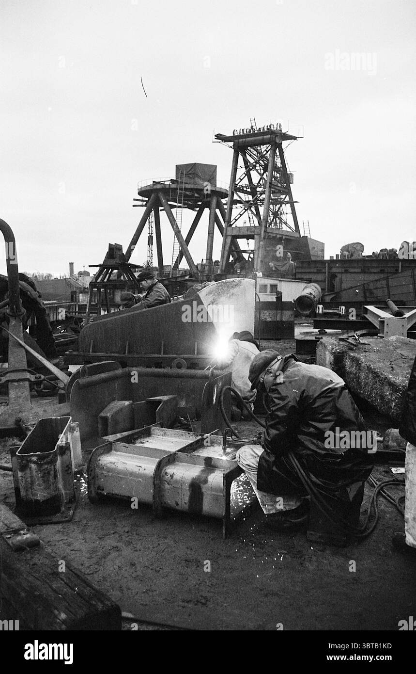 Baukraninsel im Bau bei der Gusto Werft in Schiedam Ships Work, Whizgle News, Dutch Desk, Niederlande, 1950 - 2000 am 25-11-1963. Die Abbildung zeigt diese Themen. Die Szene zeigt eine raue industrielle Umgebung, die von großen Maschinen und Strukturen dominiert wird, die auf eine schwere Arbeitsumgebung hinweisen. Im Vordergrund ist ein Arbeiter mit dem Schweißen beschäftigt, wobei der intensive Funken des Schweißbrenners den unmittelbaren Bereich mit einem hellen, weißen Fleck gegen die umgebende Dunkelheit beleuchtet. Der Arbeitnehmer in Schutzausrüstung, einschließlich einer dunklen, reflektierenden Jacke und einem Helm, Stockfoto