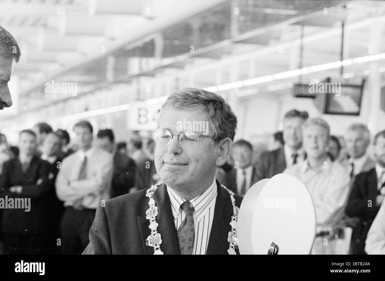 Eröffnung von Pier B Schiphol, Whizgle News, Dutch Desk, Niederlande, 1950 - 2000 am 10-08-1994. Das Bild enthält diese Themen. Die Szene fängt einen Moment in einem belebten Innenbereich ein, wahrscheinlich in einem Flughafen oder einem großen öffentlichen Veranstaltungsort, der voller Menschen ist. Im Mittelpunkt steht ein Mann mittleren Alters in einem Anzug, der ein Gefühl von Stolz und Formalität ausstrahlt. Er trägt eine Bürokette um seinen Hals, was auf eine wichtige Position hinweist, vielleicht ein lokaler Würdenträger oder Beamter. Sein Ausdruck ist zufrieden, mit einem leichten Lächeln und entspanntem Auftreten, was darauf hindeutet, dass er den Moment genießt. Um ihn herum, A Stockfoto