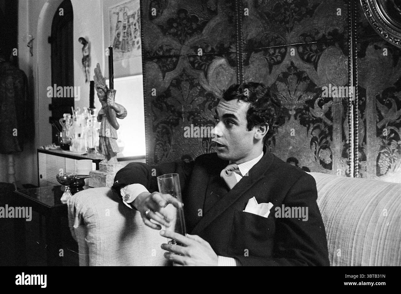 Museumsmaler Costiaan de Vries aus Laren Persons, Whizgle News, Dutch Desk, Niederlande, 1950 - 2000 am 26-07-1963. Die Abbildung zeigt diese Themen. Die Szene zeigt einen Mann, der auf einem hellen Sofa sitzt, in einem eleganten dunklen Anzug mit einem weißen Taschenquadrat gekleidet ist und ein Gefühl von Raffinesse ausstrahlt. Sein Ausdruck ist kontemplativ, mit einem Hauch von Intrigen, während er zur Seite blickt und andeutet, dass er in der Nähe nachdenkt oder jemandem zuhört. Hinter ihm ist die Wand mit einem reich gemusterten Wandteppich verziert, der einen Hauch von Vintage-Charme mit tiefen Farbtönen und komplizierten Designs verleiht. Stockfoto