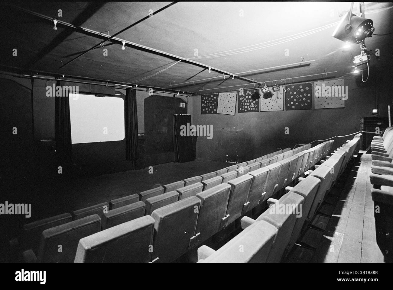Int. Cinema Pencentrale IJm. Innenausstattung, Whizgle News, Dutch Desk, Niederlande, 1950 - 2000 am 06-10-1975. Diese Themen sind in der Abbildung dargestellt. Die Szene zeigt einen schwach beleuchteten Theaterraum mit Reihen sauber ausgerichteter Sitze, die alle in einem gedämpften Rot gepolstert sind. Der Stoff scheint abgenutzt zu sein, was auf häufige Verwendung im Laufe der Zeit hindeutet. Die Sitzanordnung zeigt eine provisorische Bühne, die von schweren, dunklen Vorhängen eingerahmt wird, die an beiden Seiten hängen und teilweise das dahinter liegende verbergen. Auf der Vorderseite steht eine große weiße Leinwand bereit für die Projektion, die glatt und einladend ist. Darüber ein Spotlight Stockfoto
