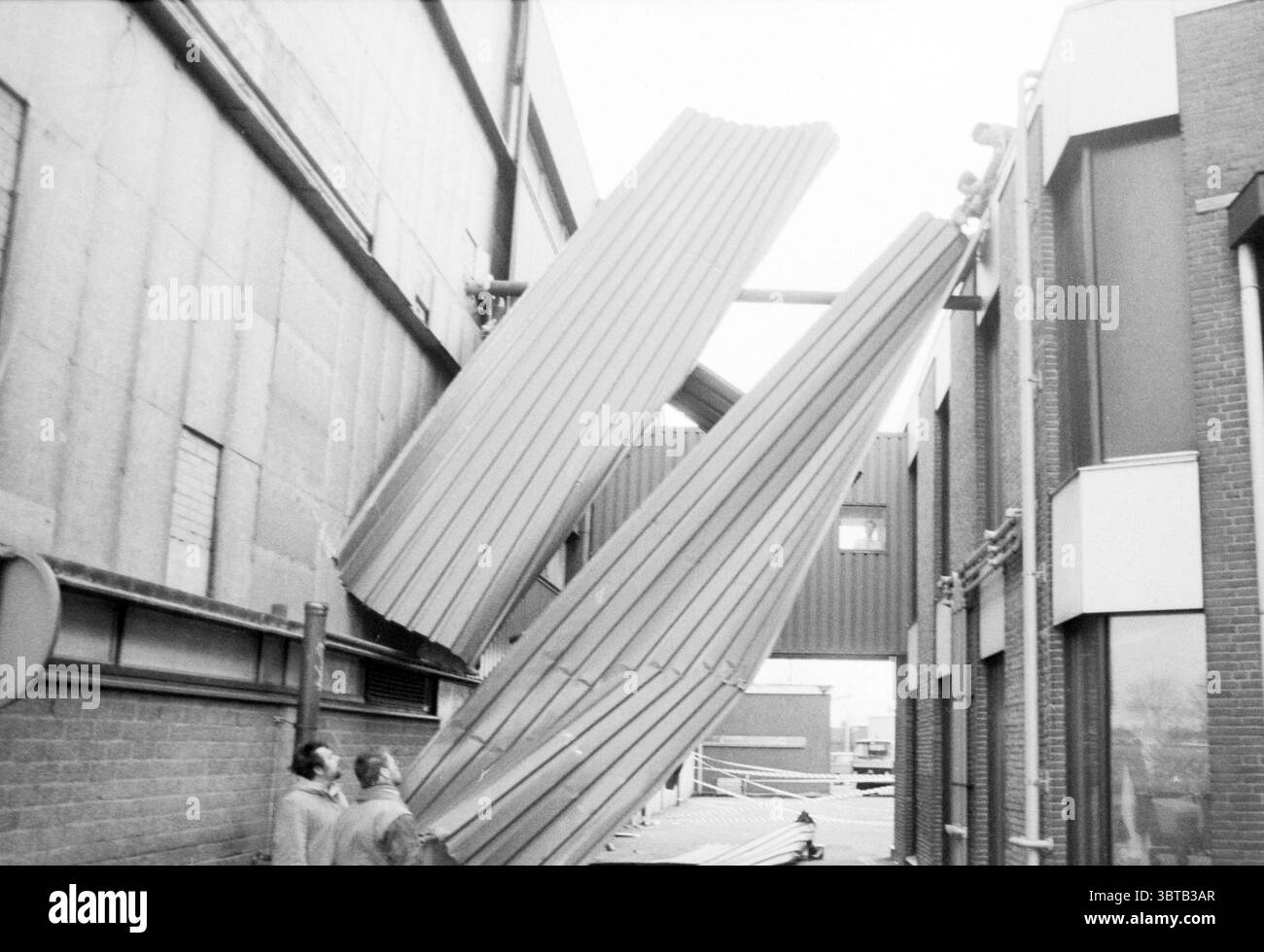 Sturmschaden verschiedene Haarlem Sturm- und Sturmschäden Haarlem the Netherlands, Whizgle News, Dutch Desk, Niederlande, 1950 - 2000 am 26-01-1990. Die Abbildung zeigt diese Themen. Die Szene fängt einen Moment ungewöhnlicher Bauten oder Industriechaos ein. Zwei große Metallbleche oder -Paneele sind stark geneigt und ragen zwischen zwei Gebäuden hervor. Die Paneele, die wie gewellt aussehen, neigen sich in einem alarmierenden Winkel, als ob sie kurz vor dem Sturz stehen oder gerade verschoben worden wären. Im Vordergrund stehen zwei Figuren, die der Struktur zugewandt sind und die Aufmerksamkeit voll auf die prekäre Positionierung der Tafeln lenken Stockfoto