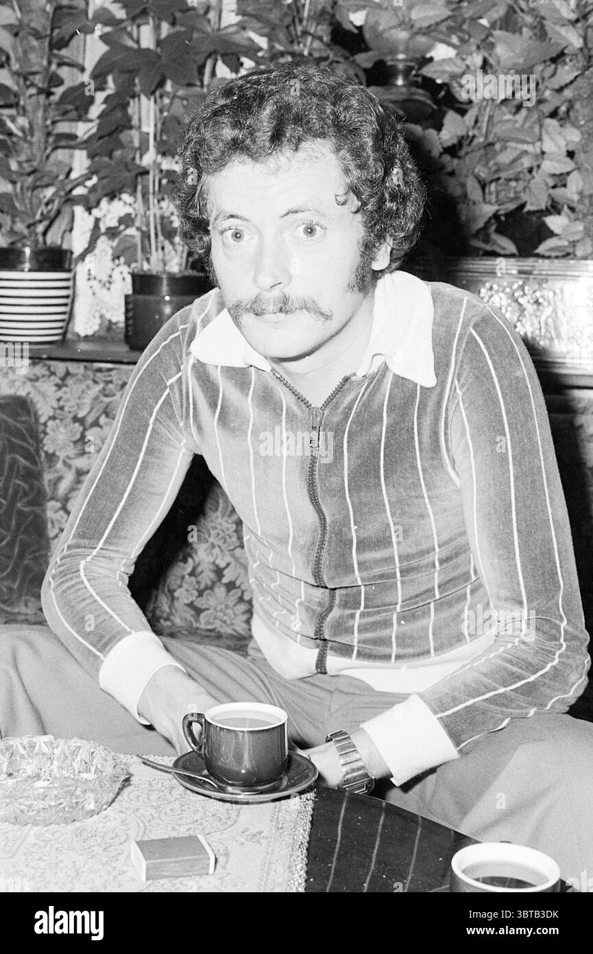 Herr Brouwer verbrannte das Auto, das verjagt (Besoekistr. H'lem) Personen Haarlem Besoekistraat Niederlande, Whizgle News, Dutch Desk, Niederlande, 1950 - 2000 am 01-11-1977. Diese Themen sind in der Abbildung dargestellt. In der Szene sitzt ein Mann auf einer gemusterten Couch, die reich strukturiert ist. Er hat eine unverwechselbare lockige Frisur und einen Schnurrbart, der seine Gesichtszüge betont. Seine Kleidung besteht aus einem Hemd mit Reißverschluss mit auffälligen, horizontalen Streifen in verschiedenen Farben, die einen auffälligen Kontrast bilden. Die Farben haben einen leicht gedämpften Ton, was auf eine Vintage-Ästhetik hindeutet. Der Mann hält einen kleinen Becher, Pres Stockfoto