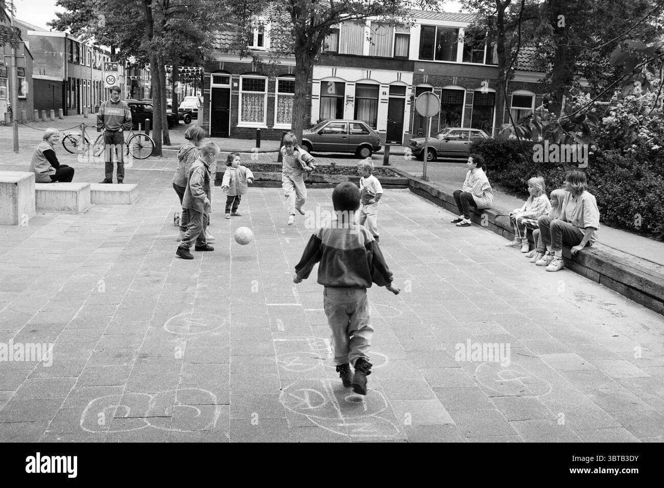 Mr. De Gelder und Spielplatz Haarlem the Netherlands, Whizgle News, Dutch Desk, Niederlande, 1950 - 2000 am 19.06.1996. Das Bild enthält diese Themen. In einer lebhaften urbanen Umgebung entfaltet sich auf einem sonnendurchfluteten Platz eine lebhafte Szene. Der Bürgersteig ist mit farbenfrohen Kreidezeichnungen und verspielten Hopscotch-Mustern verziert. Eine Gruppe von Kindern, die tatkräftig spielen, dominiert den Vordergrund. Sie zeigen eine Reihe von Ausdrücken, von Konzentration bis Freude, während sie sich in ihrem Spiel engagieren. Jedes Kind trägt eine Mischung aus Freizeitkleidung, die in verschiedenen Farben gehalten wird, die sich mit den Monochromati abheben Stockfoto
