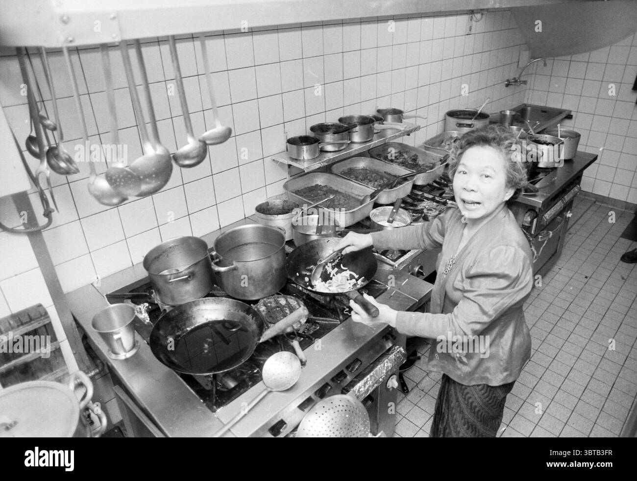 Hilda Restaurant in der Küche Haarlem Wagenweg Nederland, Whizgle News, Dutch Desk, Niederlande, 1950 - 2000 am 12-12-1991. Dies sind die Elemente im Bild. In einer belebten Küche erfährt eine ältere Frau Aufmerksamkeit, während sie Zutaten in einem großen, dunklen Topf umrührt. Sie trägt eine Schürze über einem einfachen, erdfarbenen Ensemble, ihre Haare ordentlich zurückgezogen und zeigt ihren konzentrierten Ausdruck. Die Küche ist mit einer Reihe von Metalltöpfen und Pfannen in verschiedenen Größen gefüllt, einige stapelweise mit Lebensmitteln, andere ordentlich auf Edelstahlregalen organisiert. Die Arbeitsplatten sind übersichtlich und doch zweckmäßig. Stockfoto