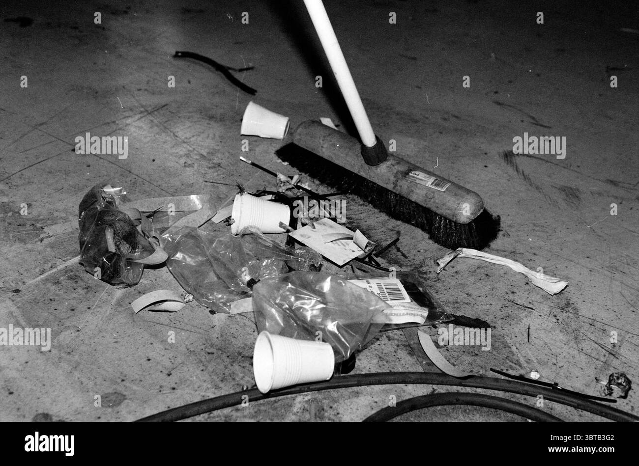 Groom and Garbage., Whizgle News, Dutch Desk, Niederlande, 1950 - 2000. Das Bild enthält diese Themen. Die Szene zeigt eine übersichtliche Umgebung, wahrscheinlich ein Arbeitsplatz oder eine Werkstatt, die durch eine flache Oberfläche mit verschiedenen Trümmern gekennzeichnet ist. In der Mitte verändert ein Besen den Fokus, dessen Borsten aktiv mit dem Boden in Berührung kommen, was darauf hindeutet, dass fortgesetzte Anstrengungen unternommen werden, um das Chaos zu beseitigen. Auf dem Boden verstreut lagen mehrere weiße Einwegbecher zufällig zwischen zerknitterten Plastikverpackungen und Papierstreifen, während sich dünne Drähte durch die Mischung schlängeln. Die gesamte Farbpalette i Stockfoto