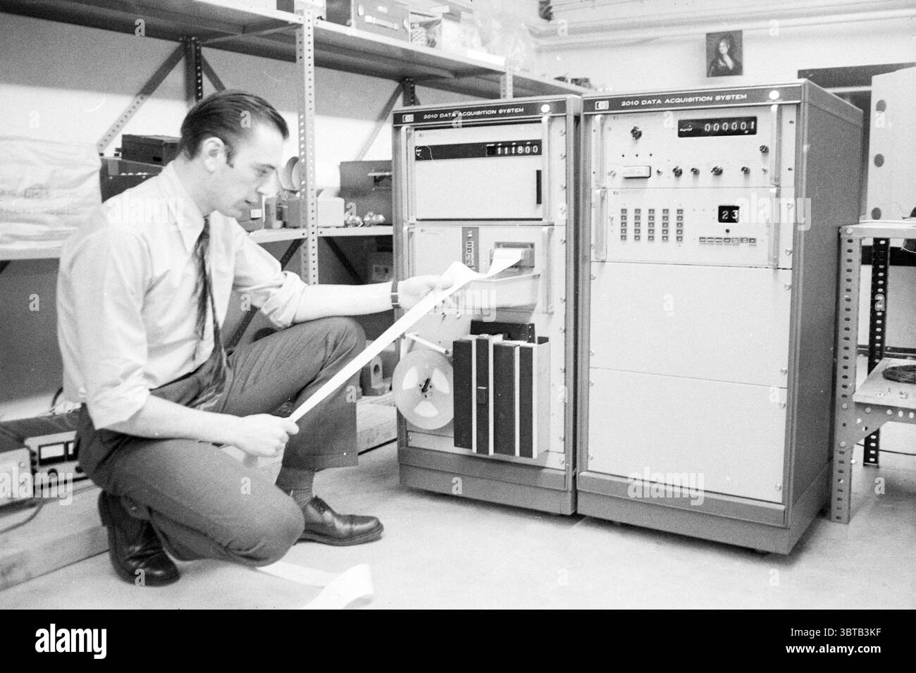 Computer für Angeln IJmuiden Maschinen und Fabriken, Whizgle News, Dutch Desk, Niederlande, 1950 - 2000 auf 14-12-1970. Dies sind die Elemente im Bild. In einer schwach beleuchteten Umgebung kniet ein Mann neben einem großen, alten Datenerfassungssystem, das sich durch seine metallisch graue Außenseite und eine Reihe von Tasten, Zifferblättern und Displays auszeichnet, die verschiedene Funktionen anzeigen. Das Gerät befindet sich auf einem stabilen, organisierten Regal aus Stahl, das einen Arbeitsbereich mit großem Sinn für Zweck bietet. Der Mann, gekleidet in einem Hemd mit Kragen und dunkler Krawatte, untersucht intensiv einen weißen Streifen PA Stockfoto