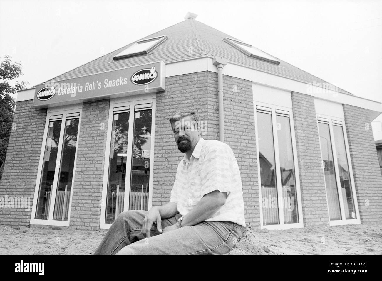 Cafeteria Rob's Snacks Snackbar Spaarndam Spaarndam, Whizgle News, Dutch Desk, Niederlande, 1950 - 2000 am 01-06-1993. Die Abbildung zeigt diese Themen. Ein Mann sitzt lässig auf einer sandigen Oberfläche vor einem einzigartigen Gebäude. Das Gebäude hat ein modernes Design, das sich durch sein kantiges Dach und eine markante Glasfassade auszeichnet. Die Wände sind aus hellen Ziegeln gefertigt, was der gesamten Ästhetik einen rustikalen Touch verleiht. Mehrere große Fenster reflektieren das Tageslicht und schaffen eine helle und einladende Atmosphäre. Der Mann mit Bart und kurzen Haaren trägt ein gemustertes, locker geschnittenes Hemd und strahlt ein entspanntes aus Stockfoto