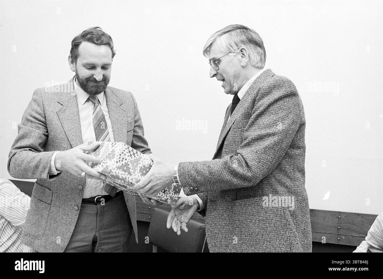 Abschied Cor Hoogland IJmuiden (Pilotendienst) Pilotenpiloten IJmuiden Niederlande, Whizgle News, Dutch Desk, Niederlande, 1950 - 2000 am 06-04-1982. Das Bild enthält diese Themen. Die Szene zeigt zwei Männer, die in einem Moment des Geschenks verwickelt sind, beide in formeller Kleidung. Ein Mann mit einem Bart und einem freundlichen Ausdruck überreicht ein hell gewickeltes Geschenk mit einem komplizierten Muster, das in leuchtenden Farben und geometrischen Formen verziert ist. Der andere Mann trägt eine Brille und einen Anzug mit einem dezenten karierten Muster und zeigt einen Blick der Wertschätzung und Neugier, während er die Prägung empfängt Stockfoto