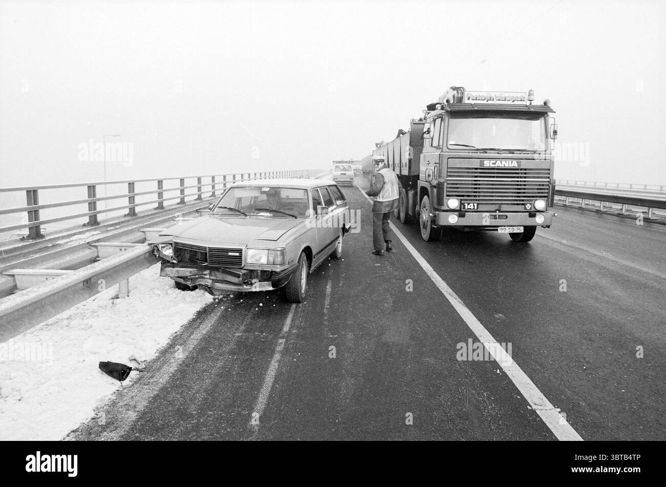 Kollision von Auto und Polizeimotorrad auf der Autobahn, Whizgle News, Dutch Desk, Niederlande, 1950 - 2000 am 06-01-1986. Das Bild enthält diese Themen. Die Szene fängt eine winterliche Autobahn ein, wo ein Gefühl von Strenge die visuelle Atmosphäre durchdringt. Ein grauweißer Nebel verwischt den Horizont und trägt zu einer düsteren Stimmung bei. Auf der rechten Seite erhebt sich ein großer Lkw, dessen metallische Karosserie leicht im gedämpften Licht leuchtet, während die Kabine mit reflektierenden Streifen verziert ist, die ins Auge fallen. Im Vordergrund steht ein kleineres Auto, das leicht beschädigt und mit einem Zerknitter vorn verkrümmt ist, in der Nähe von t Stockfoto