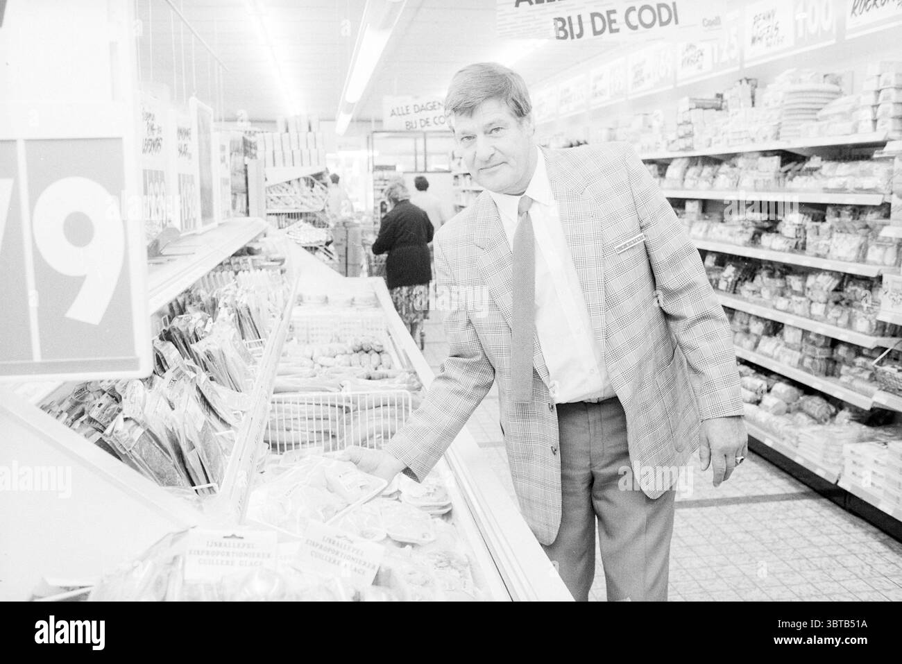Herr Sintenie CODI Santpoort Supermarket Santpoort, Whizgle News, Dutch Desk, Niederlande, 1950 - 2000 am 12.06.1987. Dies sind die Themen im Bild. In einem belebten Lebensmittelgeschäft steht ein Mann in der Nähe einer Ausstellung verschiedener Fleischprodukte. Er trägt einen hellen, gemusterten Blazer und ein knuspriges weißes Hemd mit Krawatte, was ihm ein poliertes Aussehen verleiht. Der Mann hat eine selbstbewusste Haltung, lehnt sich leicht nach vorne, während er die Gegenstände vor ihm inspiziert und einen fokussierten Ausdruck zeigt. Um ihn herum sind die Regale mit verschiedenen verpackten Waren und Fleisch gesäumt, wodurch ein Sen entsteht Stockfoto